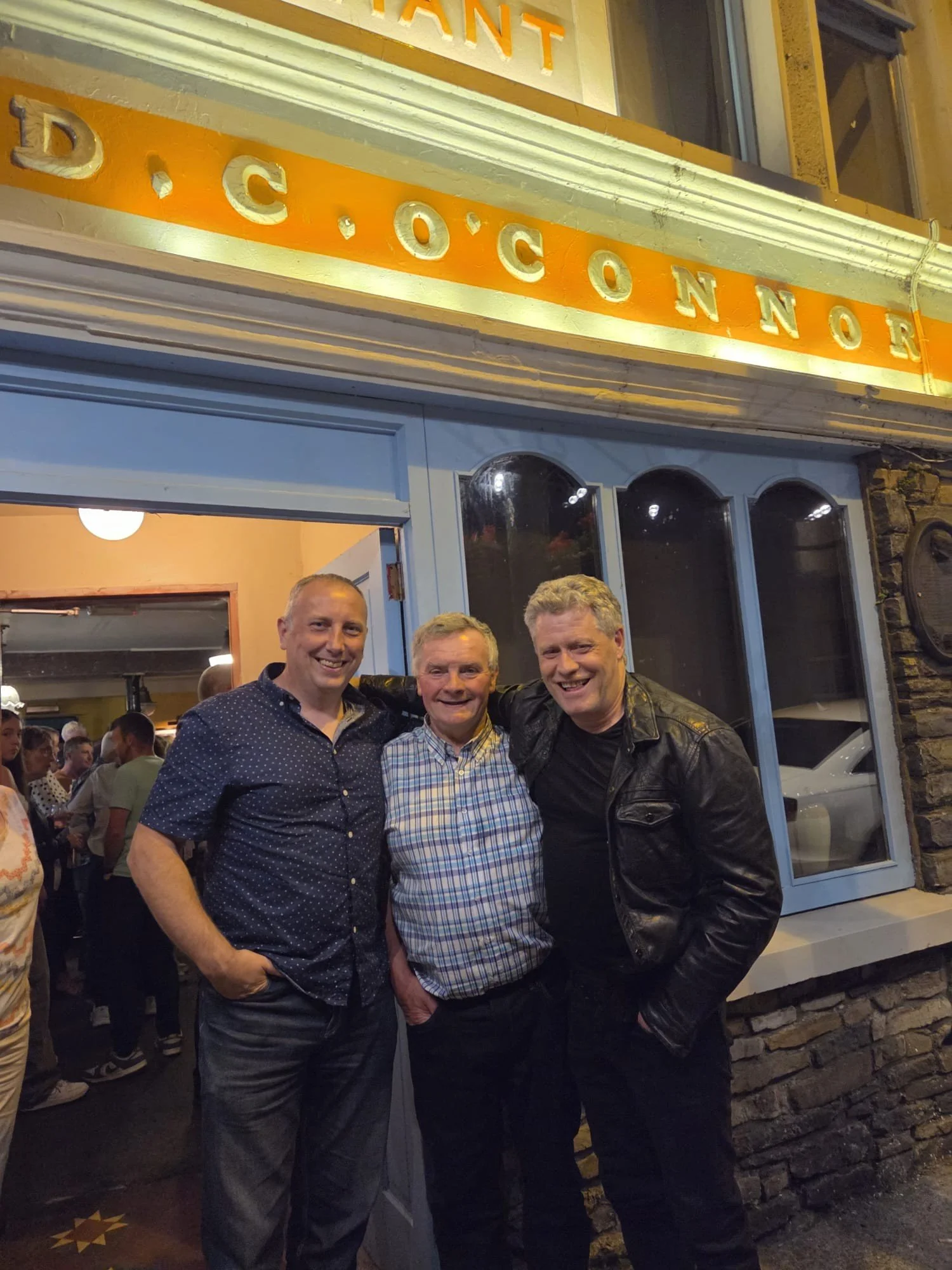 Four men are standing outside a restaurant or bar, smiling for the camera. Behind them is a sign that reads 'DACONNOR' and a large window, with other people visible inside.