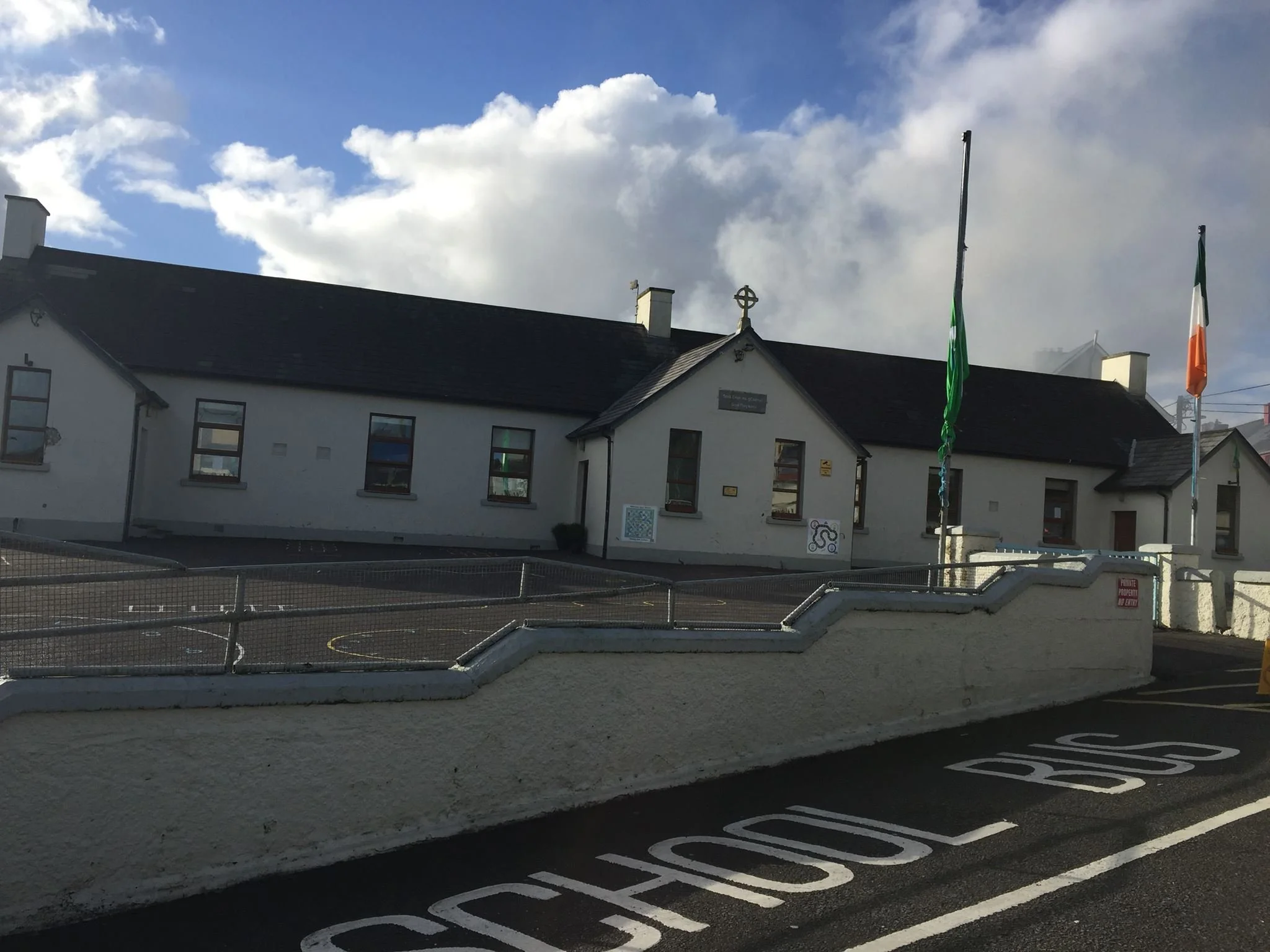 A church with a black roof and white walls, three flags (green, white, and orange), and a cross on the roof, with a partly cloudy sky in the background. There is a small parking lot and a low fence in front.