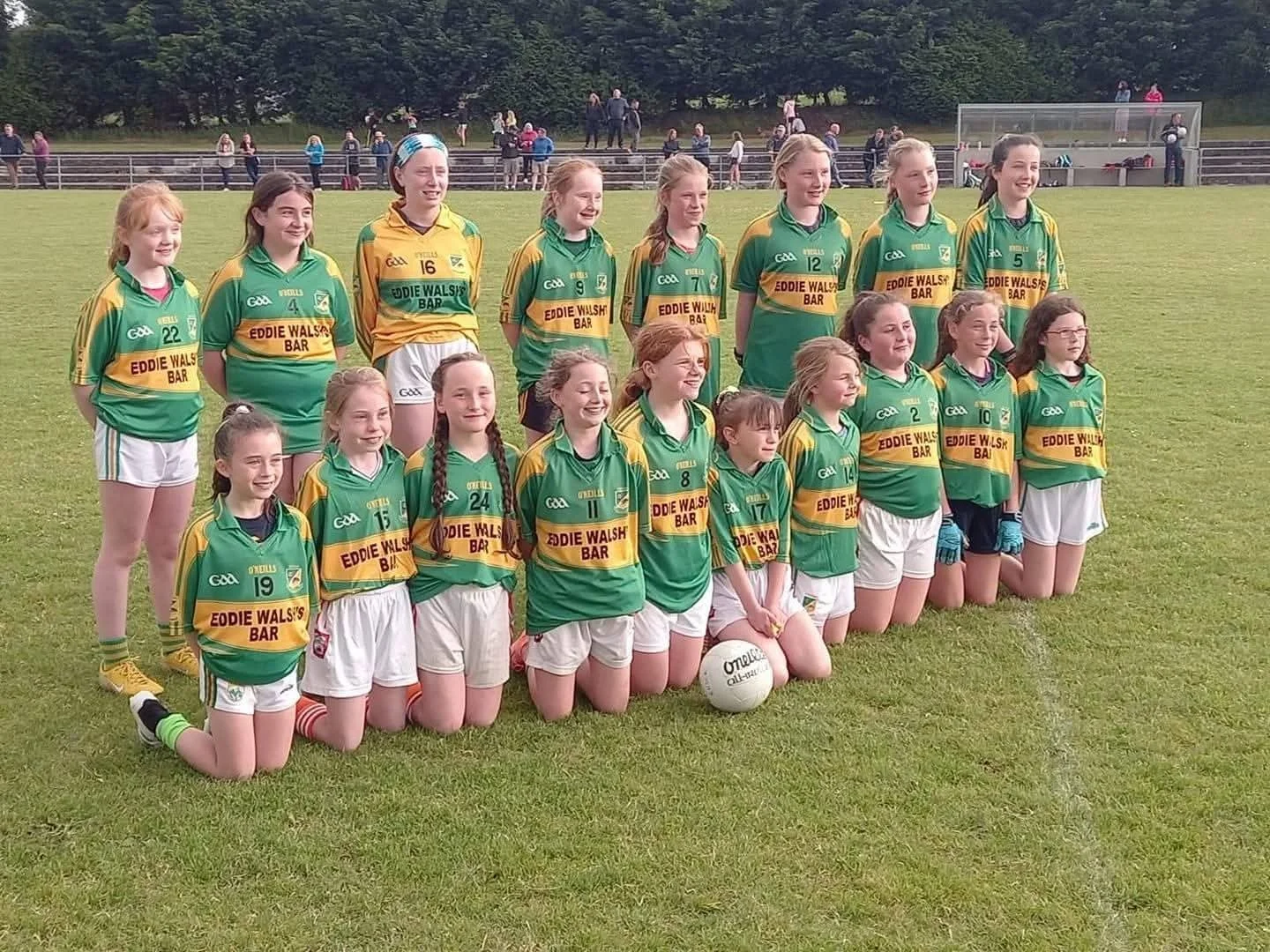 A girls' sports team on a grass field, wearing green and yellow jerseys with the sponsor 'Eddie Walsh's Bar,' some kneeling and others standing, with a soccer ball at their feet, and spectators in the background.