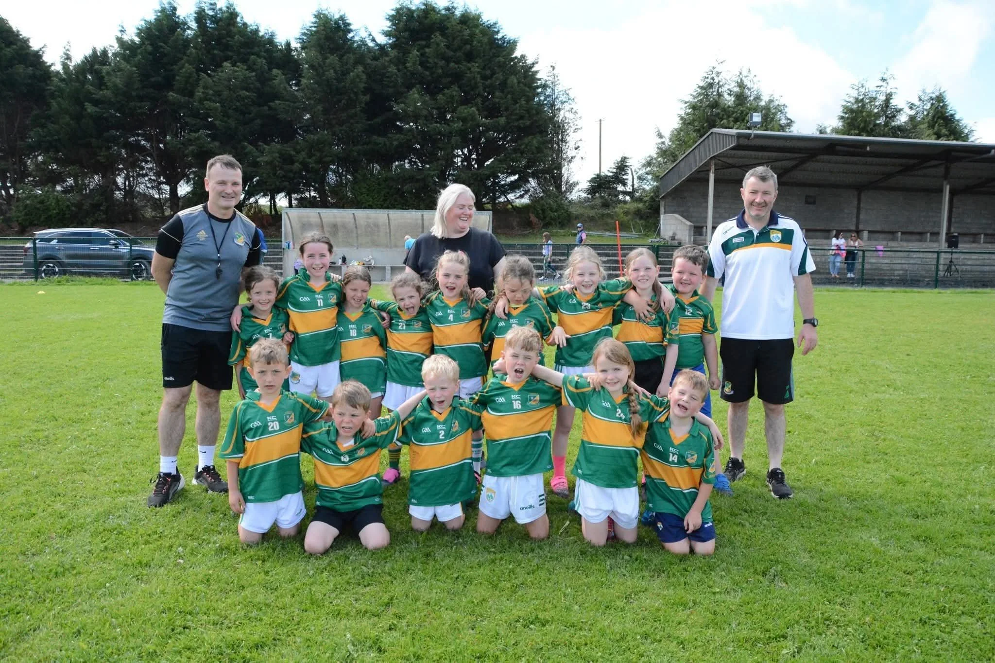 A youth sports team with adult coaches, posing on a grassy field after a game. The team wears green and yellow uniforms, and the coaches wear sports shirts. Some children are kneeling in front, and others are standing behind, smiling.