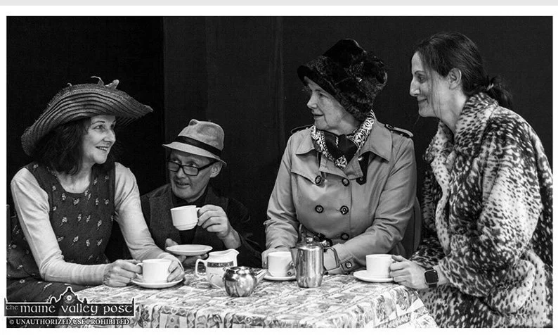 Four women sitting at a table with tea cups, a teapot, and a sugar bowl, smiling and talking. They are dressed in casual and vintage-inspired clothing, with one woman wearing a wide-brimmed hat and another in a patterned coat.