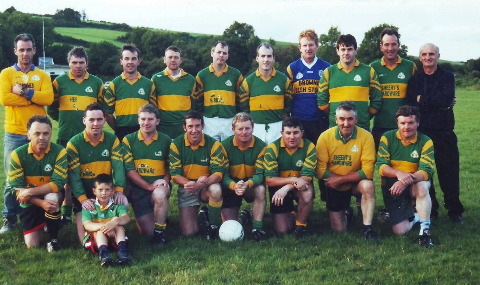 A group of adult men and one boy in green and yellow sports uniforms gathered on a grassy field, posing for a team photo with a soccer ball in front, with hills and trees in the background.