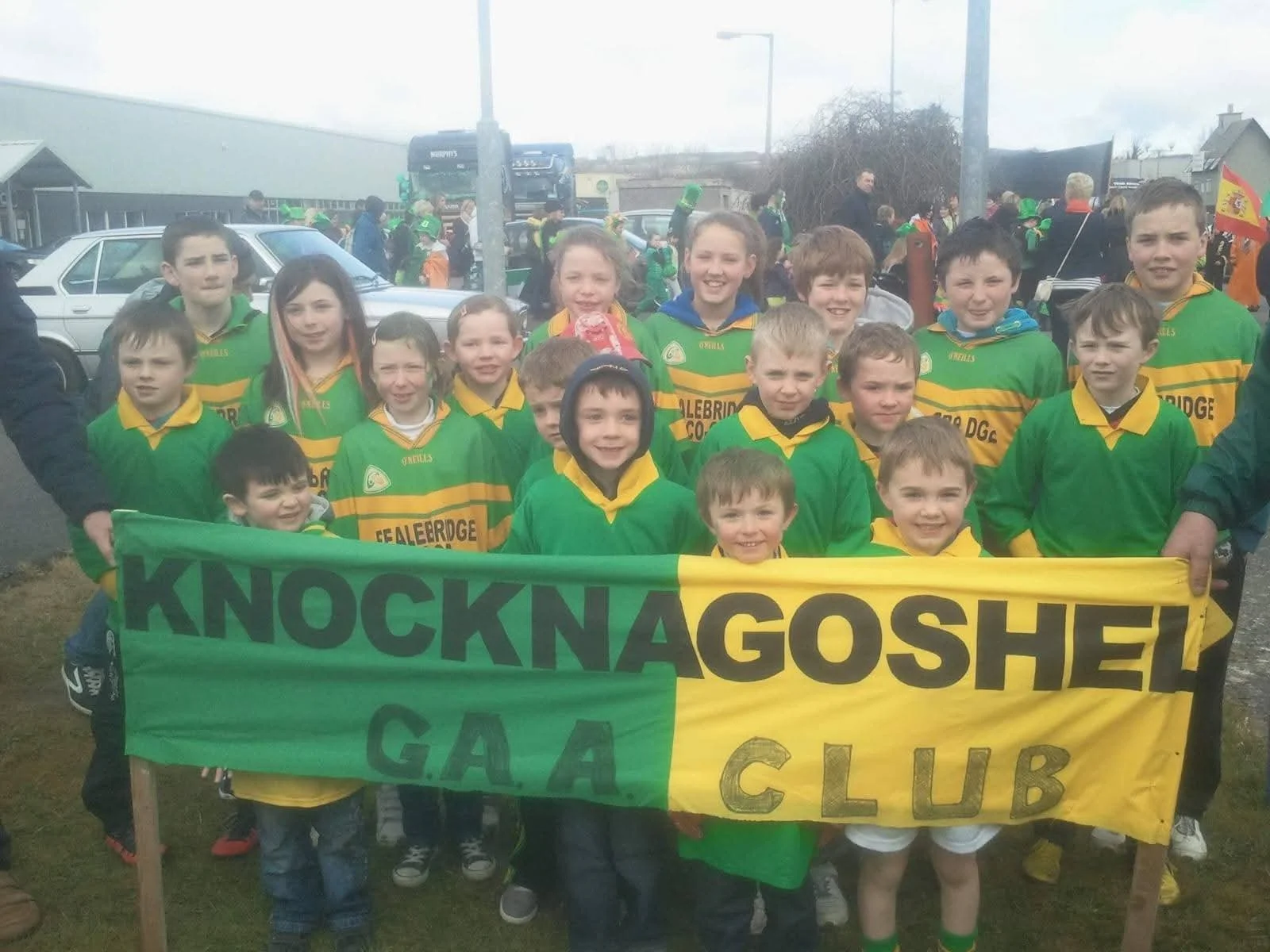 A group of children and teenagers wearing green and yellow sports jerseys holding a green and yellow banner that reads 'KNOCKNAGOSHEL GAA CLUB'. They are outdoors, possibly at a sports event or parade.