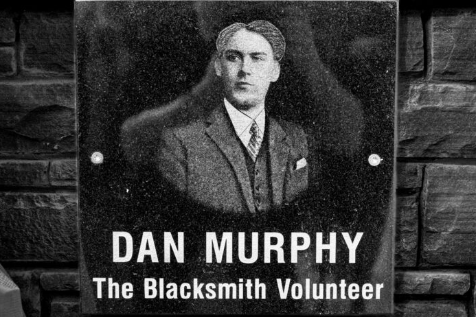 Black and white photograph of a man in a suit with the text "DAN MURPHY, The Blacksmith Volunteer" below the photo.