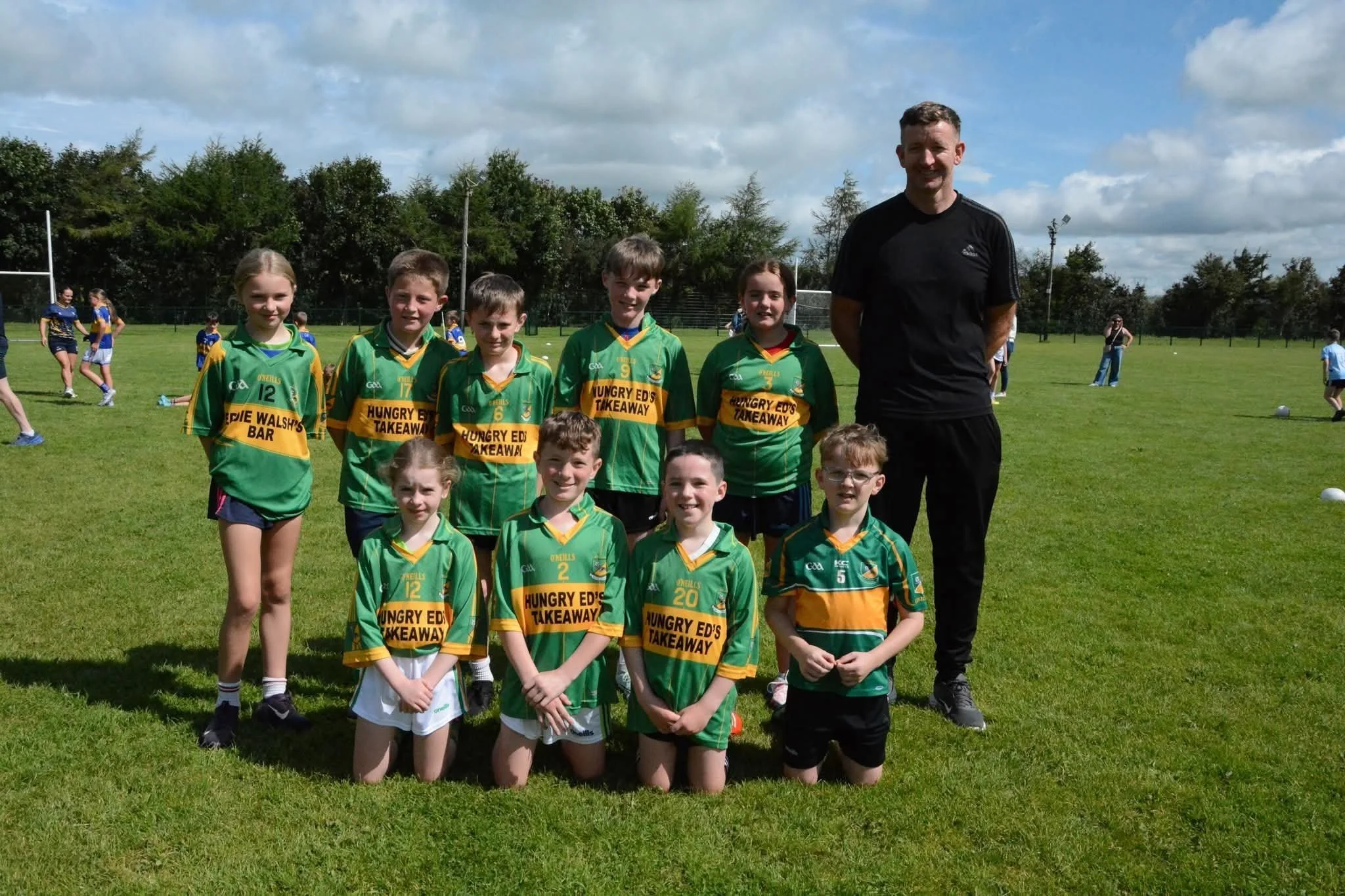 A group of children and a coach posing for a photo on a grassy field during a sports event, with children wearing green and yellow sports uniforms and the coach in black.