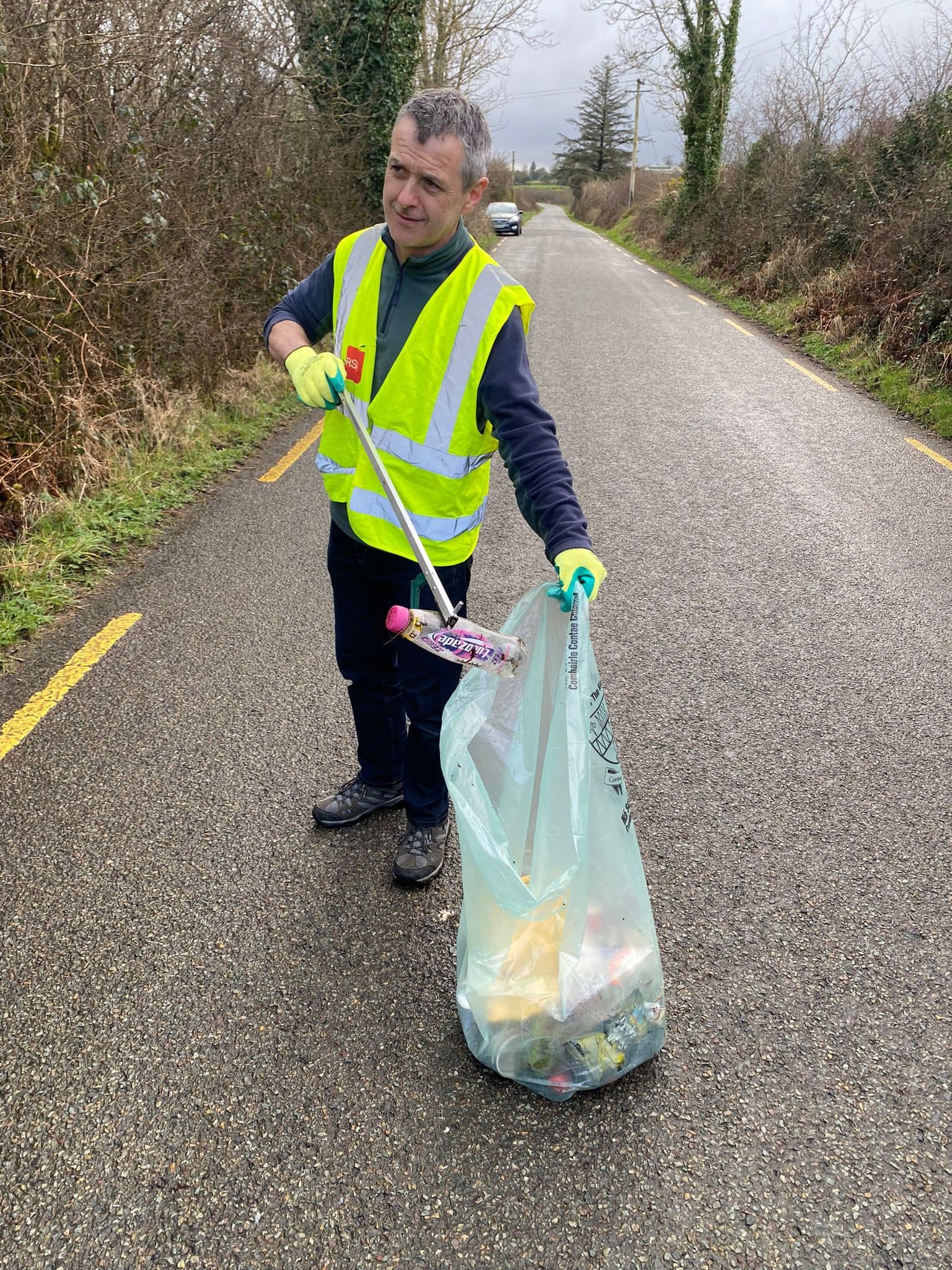 A man in a high-visibility vest and gloves holding a trash grabber and a large trash bag filled with collected litter on the side of a rural road.