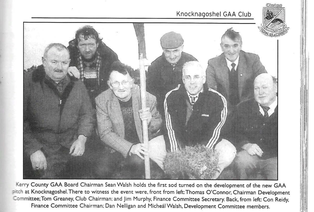 Group of seven men sitting and standing, with one holding a sheaf of straw and a long stick, in front of a banner reading "Knocknagoshel GAA Club."