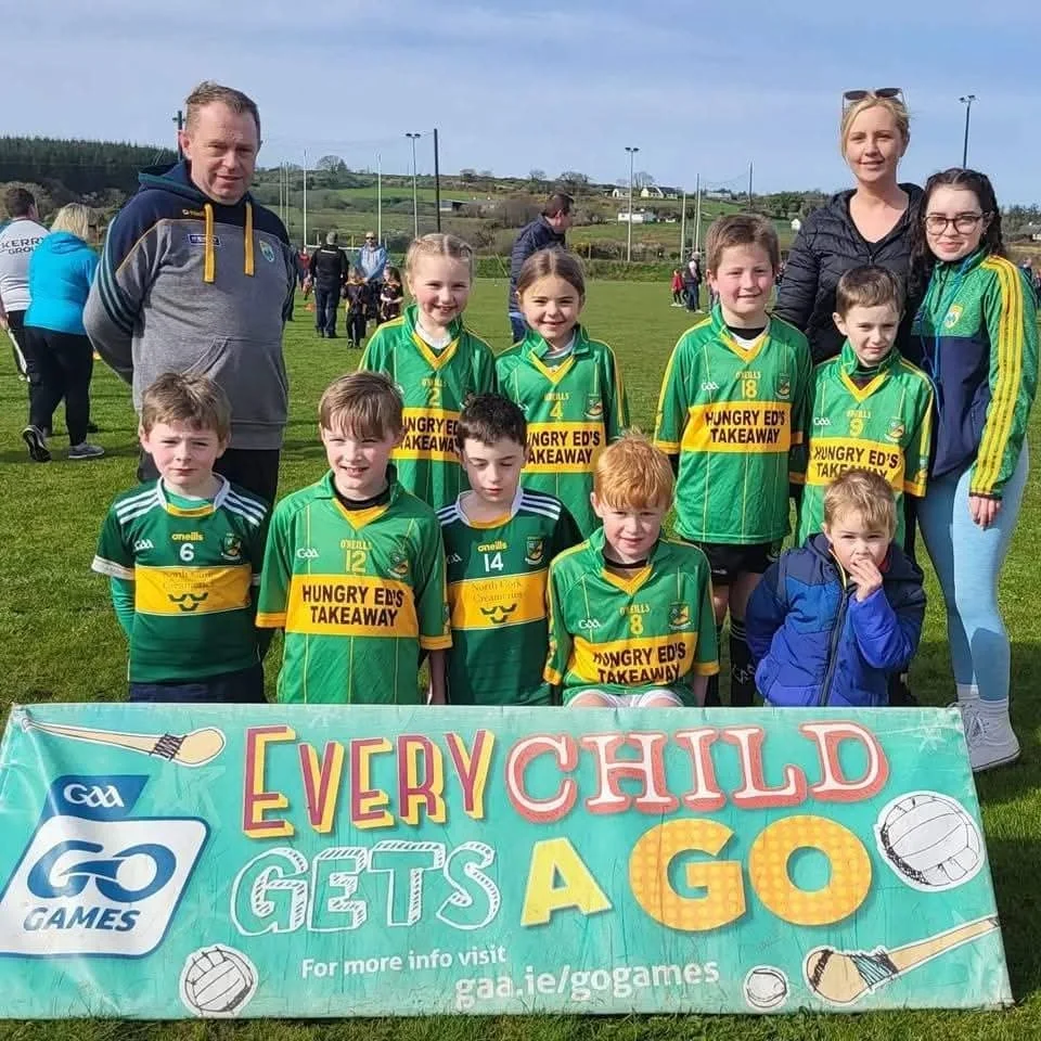 Group photo of young children in green sports uniforms with two adult women, on a grassy field, holding a banner for GAA GO GAMES, during a sports event.