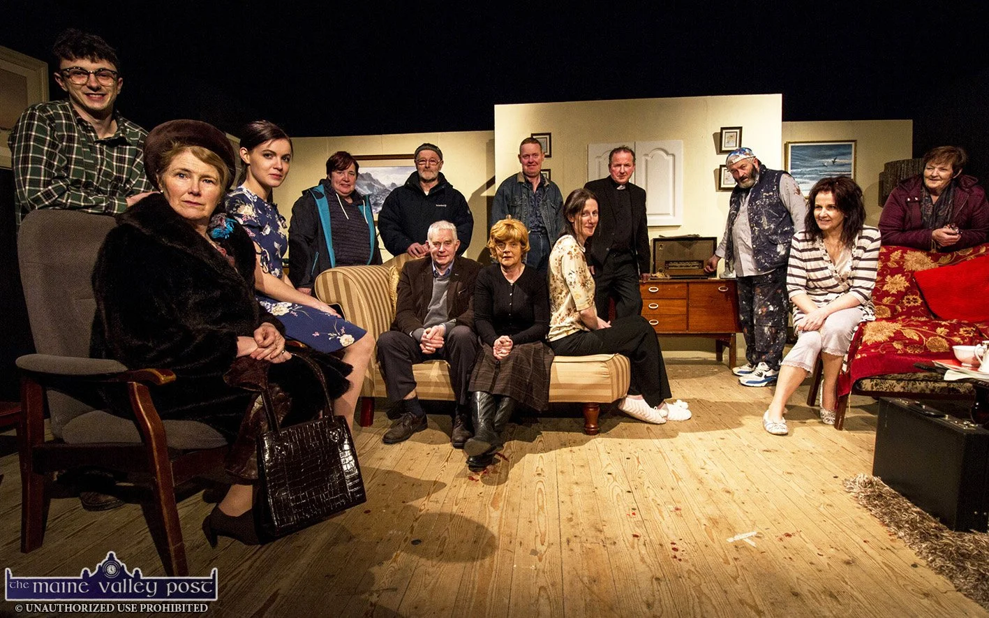 Group of people on stage in a play, posing for a photo in a living room scene with furniture and wall decorations.