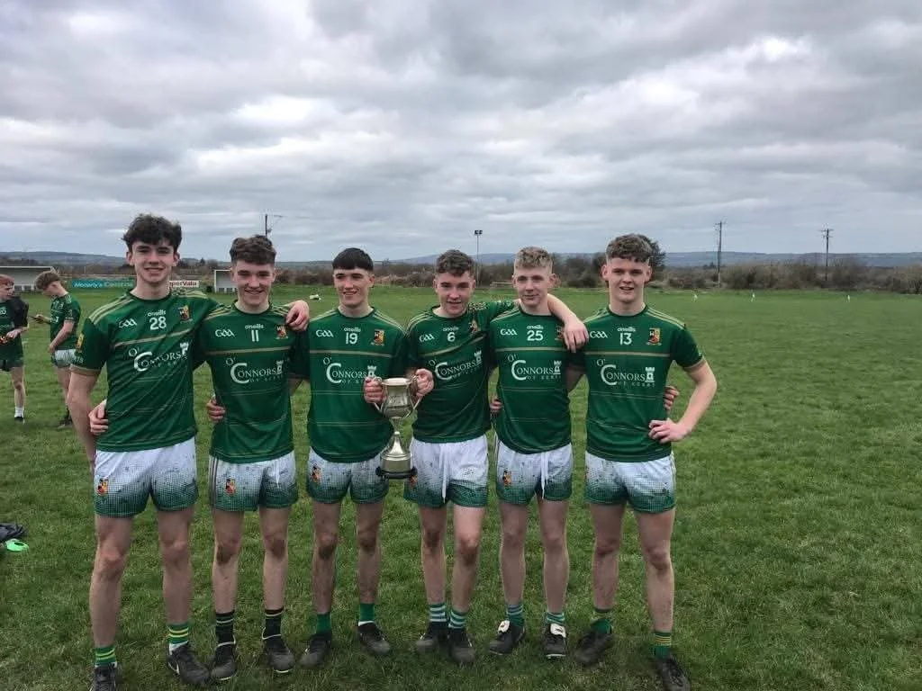 Group of seven young male athletes in green sports uniforms standing on a grassy field, holding a trophy, celebrating after a game.