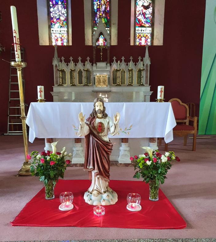 Religious altar inside a church with a statue of Jesus Christ in the foreground, decorated with flowers, candles, and a red cloth, with stained glass windows in the background.