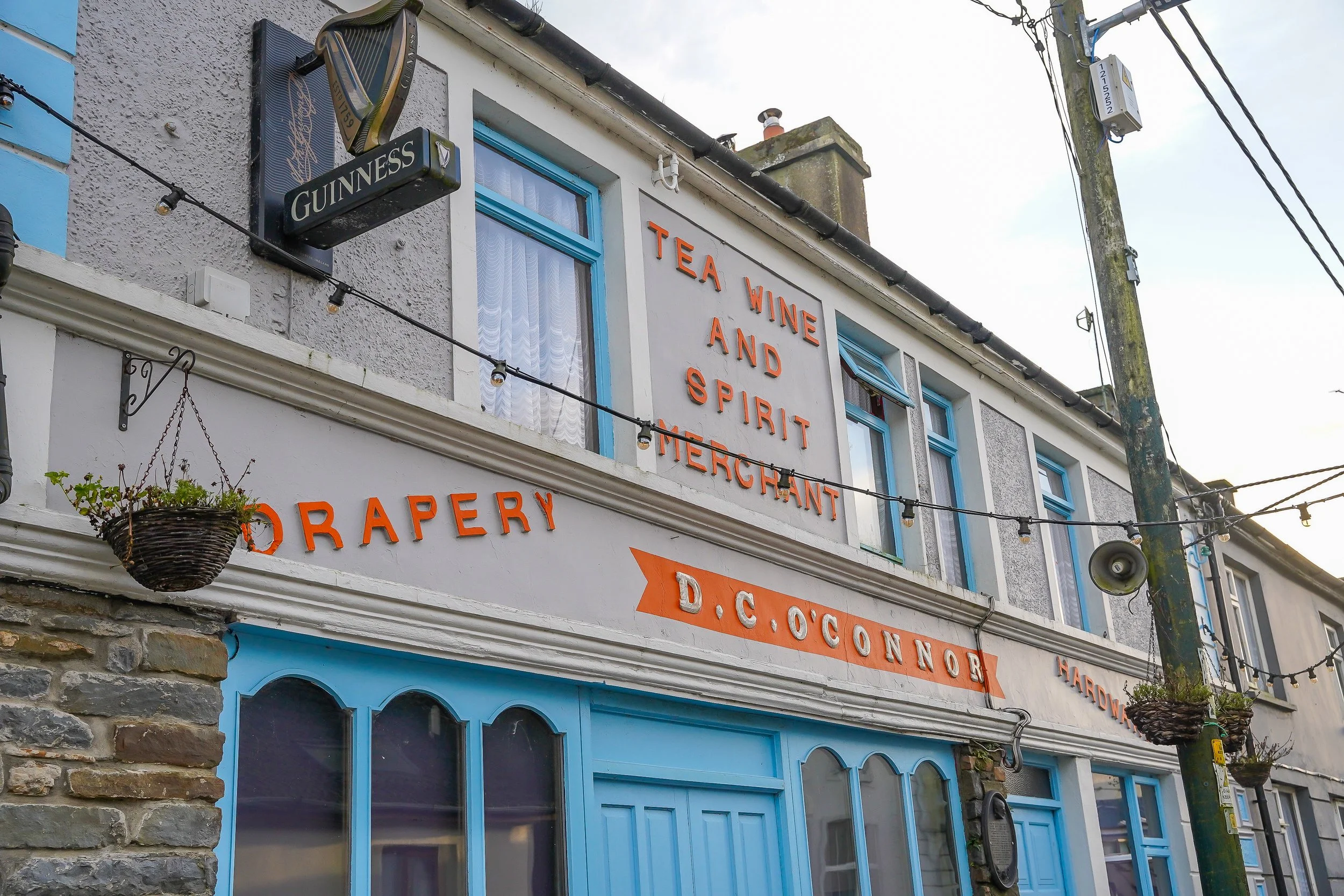 A storefront with signs that read 'Tea, Wine, and Spirit Merchant' and 'D.C. Confectionery.' The building has blue windows and doors, hanging flower baskets, a Guinness sign, and a stone facade at the corner.
