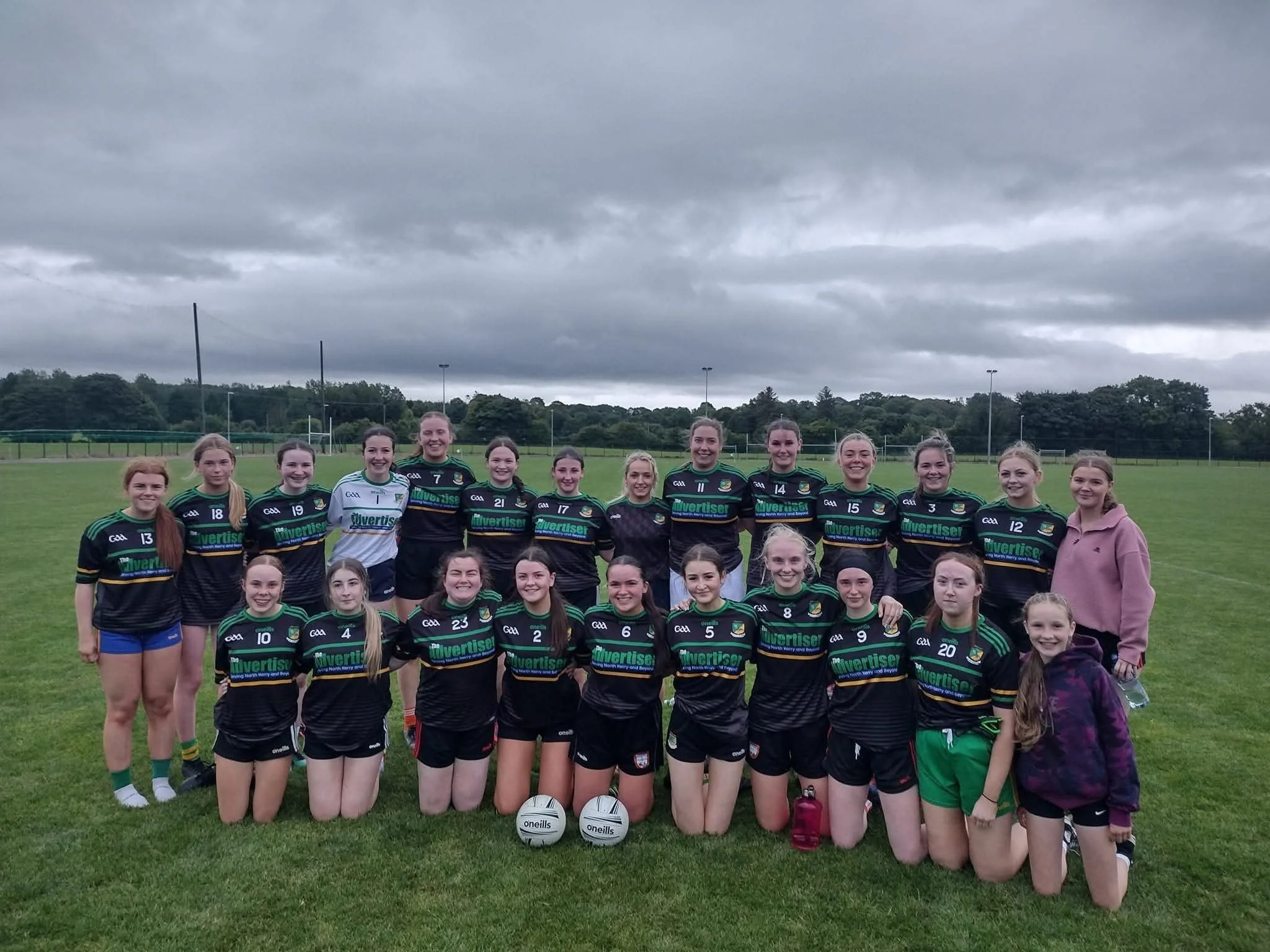 Group of young female athletes in matching black and green team uniforms, posing for a photo on a green field with overcast skies, with some kneeling and others standing, a soccer ball in front.