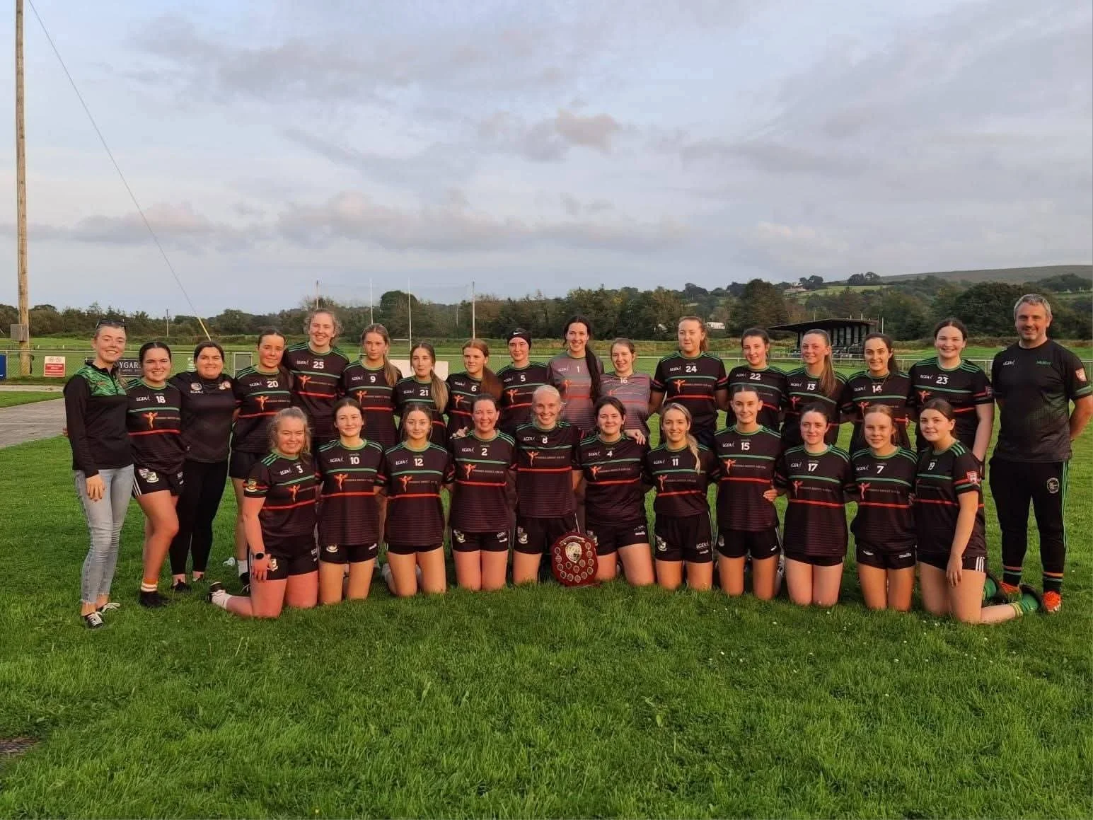 A women's sports team of 21 players and 2 coaches standing on a grassy field outdoors, wearing black team uniforms with green accents, posing for a photo in front of a rugby field with goals and distant hills.