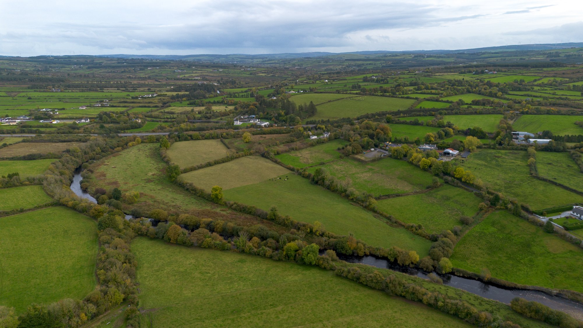 Aerial view of a lush green rural landscape with fields, small roads, scattered buildings, and a winding river under a cloudy sky.