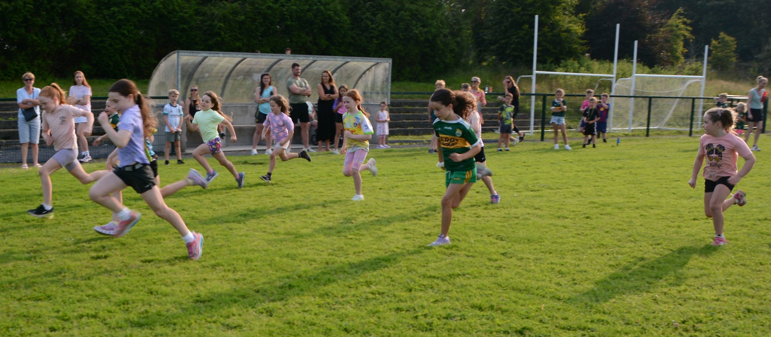 Children running and playing on a grassy field, with spectators watching, near a sports goal post.