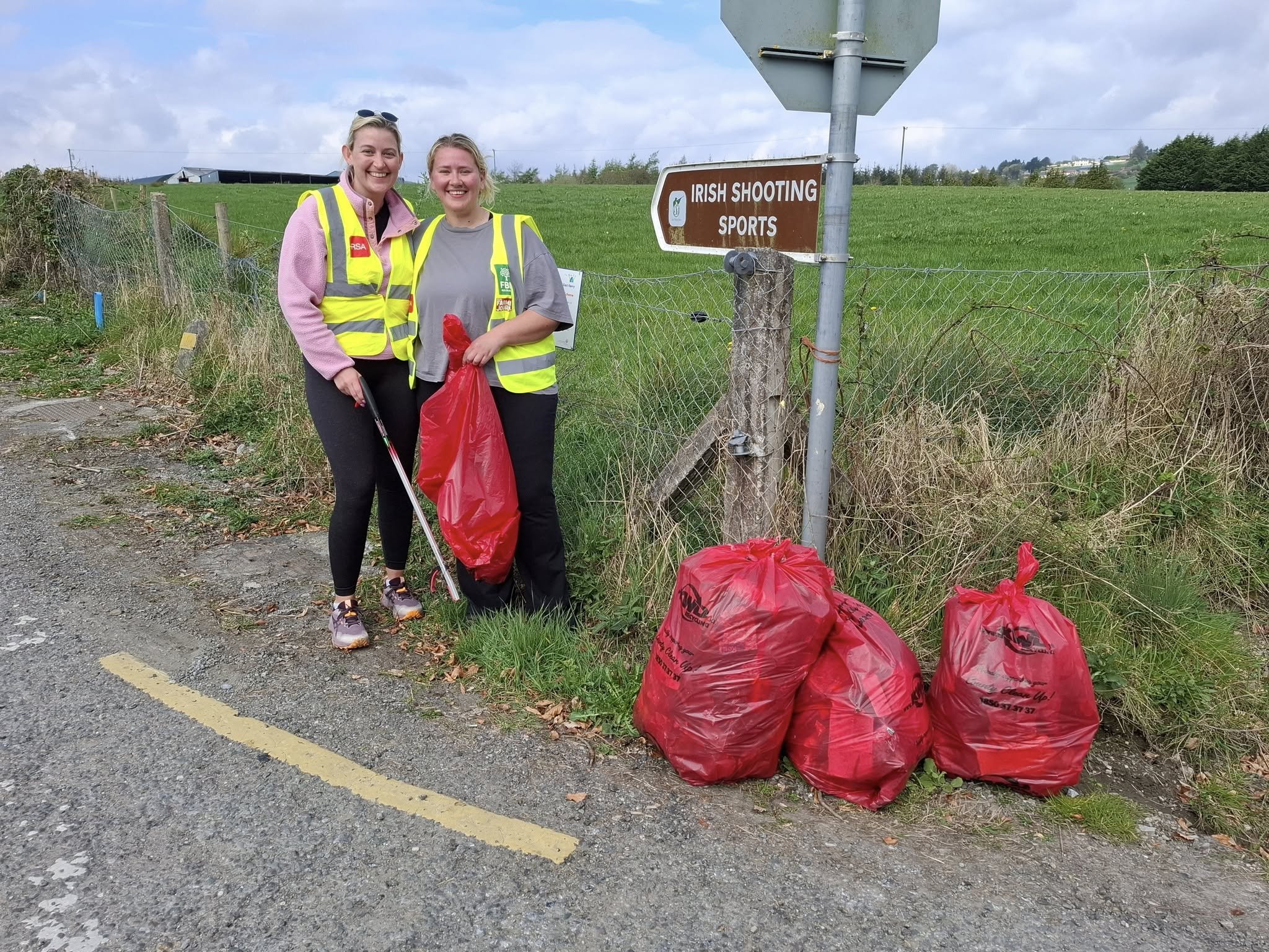 Two women standing outdoors, smiling, holding trash bags and a litter picker near a fence and a sign that reads 'Irish Shooting Sports,' with green fields and cloudy sky in the background.