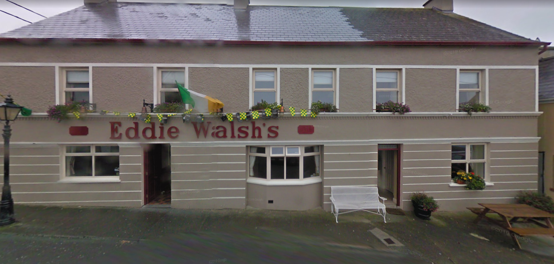 A building with a sign that reads 'Eddie Walsh's' on the front, decorated with Irish flags and bunting. The building has two floors with windows and flower boxes, a bay window, a white bench outside, and a picnic table on the sidewalk.