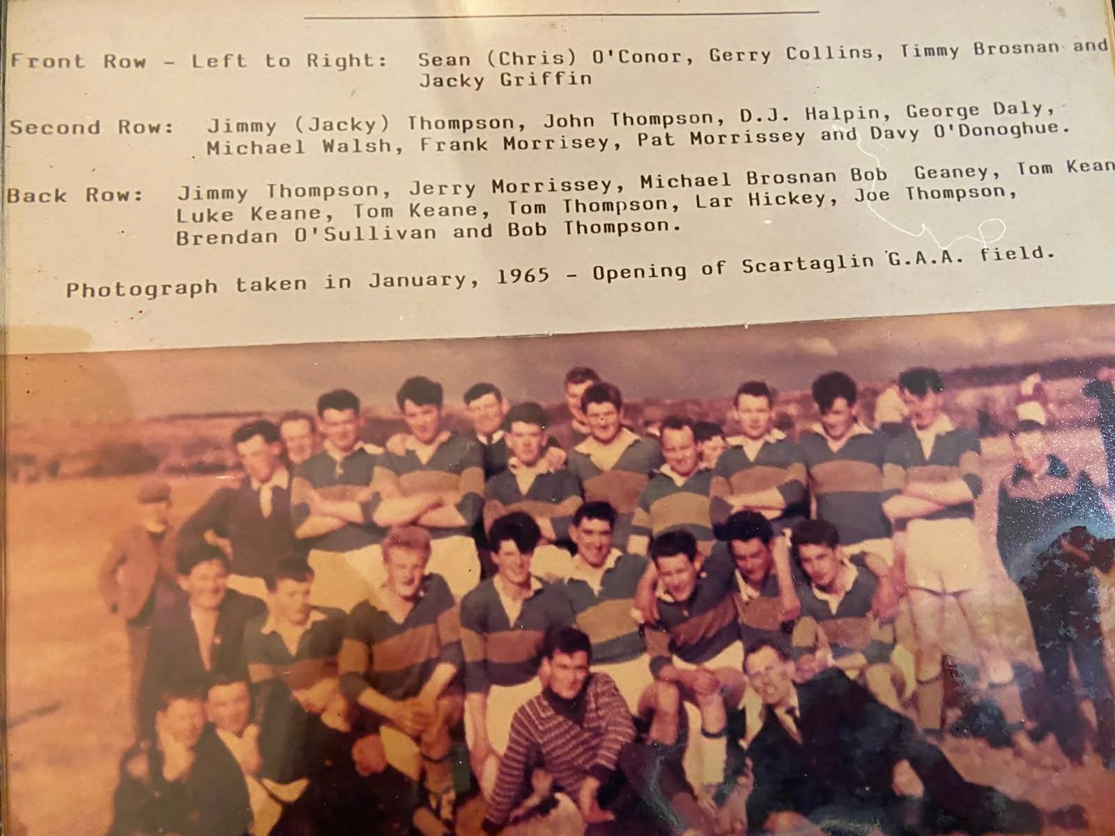 A vintage team photograph of young men in rugby uniforms, posing outdoors in a field with open landscape in the background, taken in January 1965 during the opening of Scartaglin G.A.A. field.