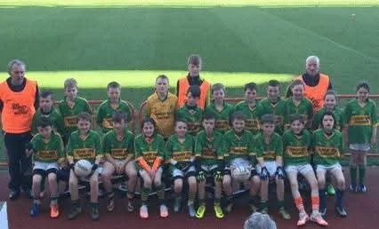 Group of young soccer players in green uniforms with orange vests, posing for a team photo on a field, with some holding soccer balls, and two coaches standing on sides.