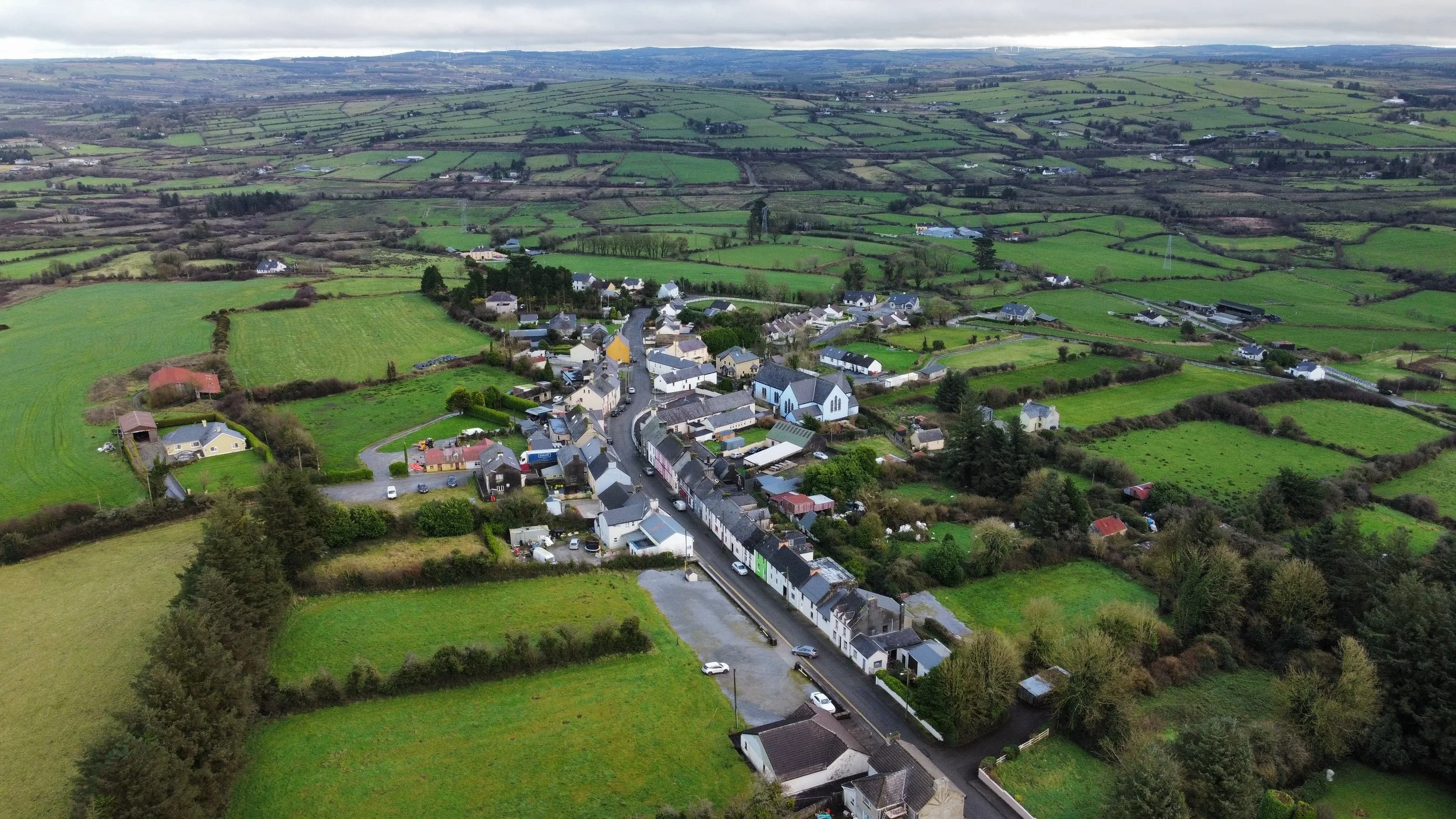 Aerial view of a small town surrounded by green fields and patchwork farmland under cloudy sky.