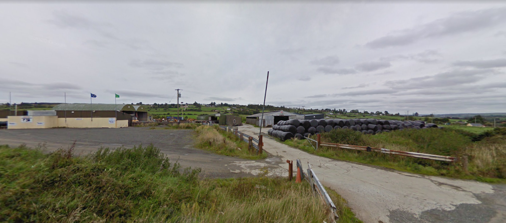 Rural industrial area with storage buildings, large black pipes stored outdoors, and rolling green hills in the background under cloudy sky.