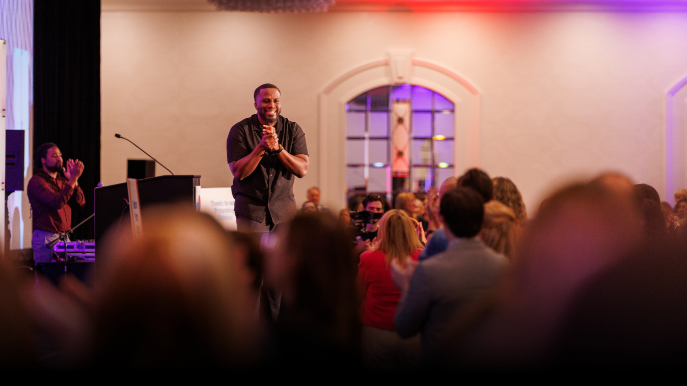 A man standing on a stage, smiling and clapping, in front of an audience during a presentation or event.
