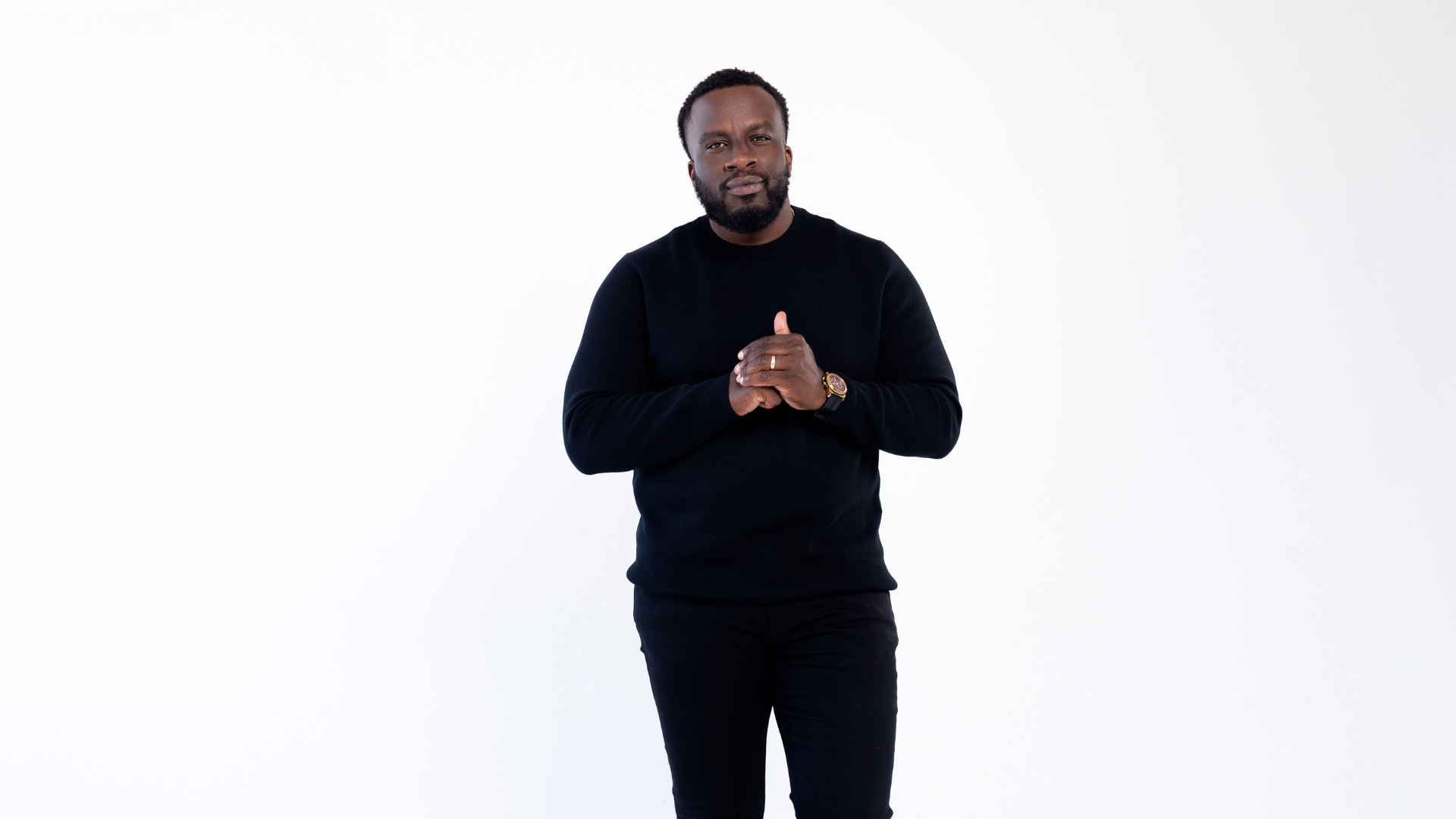 A man with dark skin, a beard, and short curly hair, standing against a plain white background. He is wearing a black long-sleeve sweater, black pants, a watch, and a ring on his left hand. He has a serious expression, with hands clasped together near his chest.