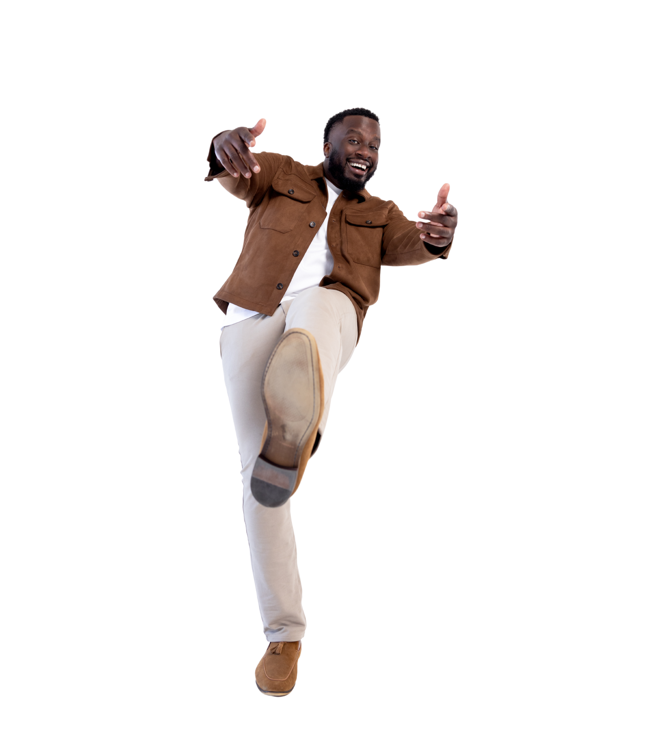 A smiling man in a brown jacket and beige pants pointing towards the camera with his left hand and lifting his right foot forward in a playful pose.