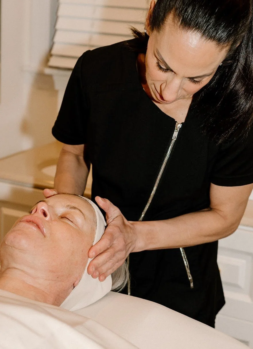Liz Clapsis giving a facial massage to an older woman lying down with a facial mask. The woman is wearing a black top and has dark hair, while the older woman has light-colored hair and a headband.