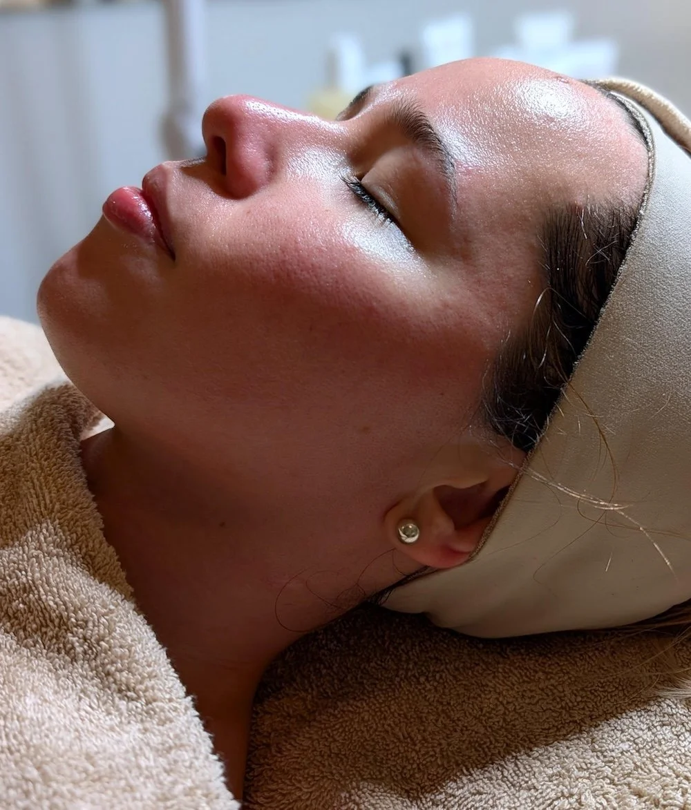 A woman with smooth skin receiving a facial treatment, lying with her eyes closed, wearing a headband and pearl earring.