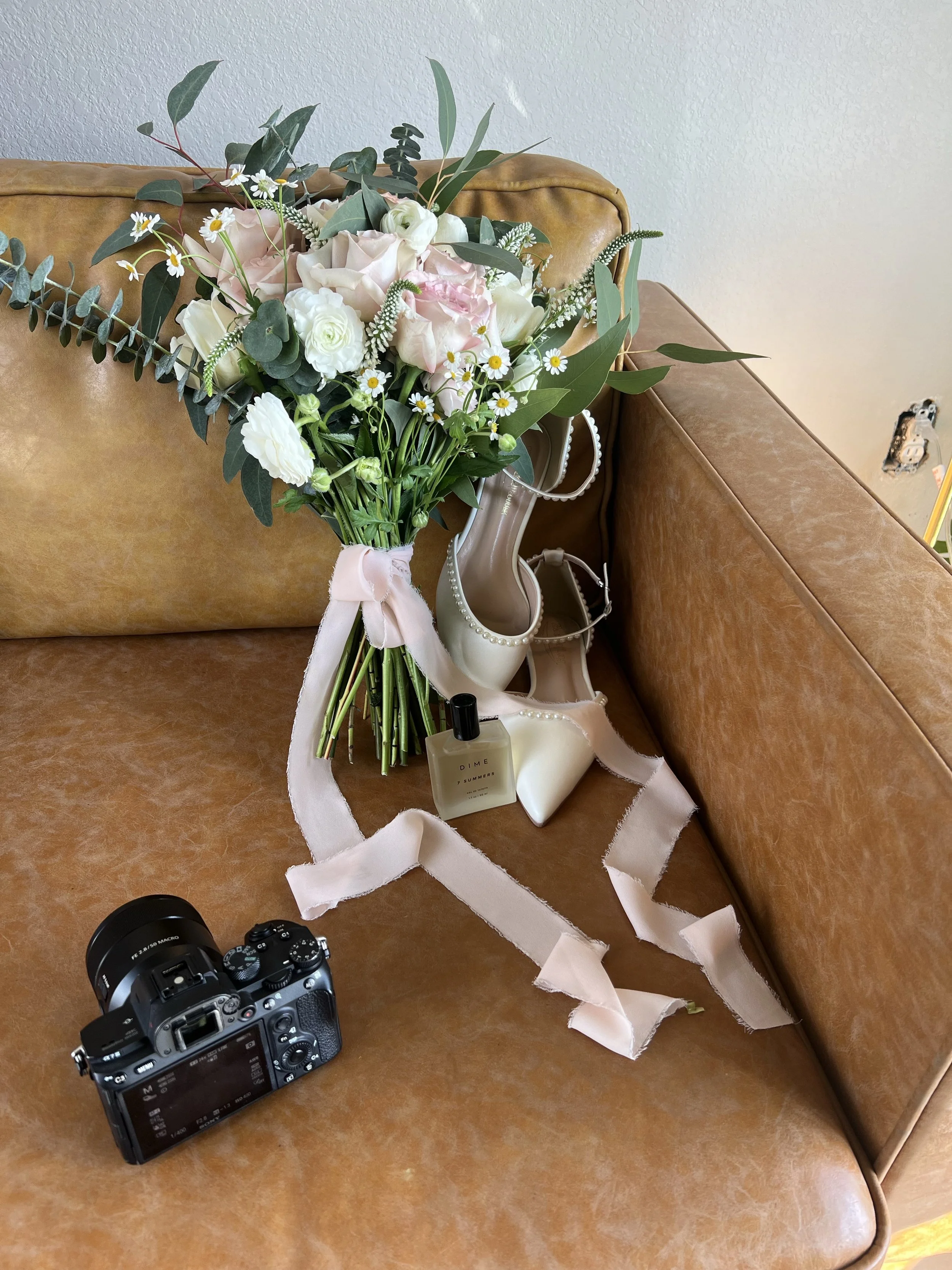 A floral bouquet with white and pink flowers, greenery, and a pink ribbon tied around the stems, placed on a brown leather sofa. There is a pair of white high-heeled shoes with pearl accents, a bottle of perfume, a camera, and a strip of pink fabric on the sofa.