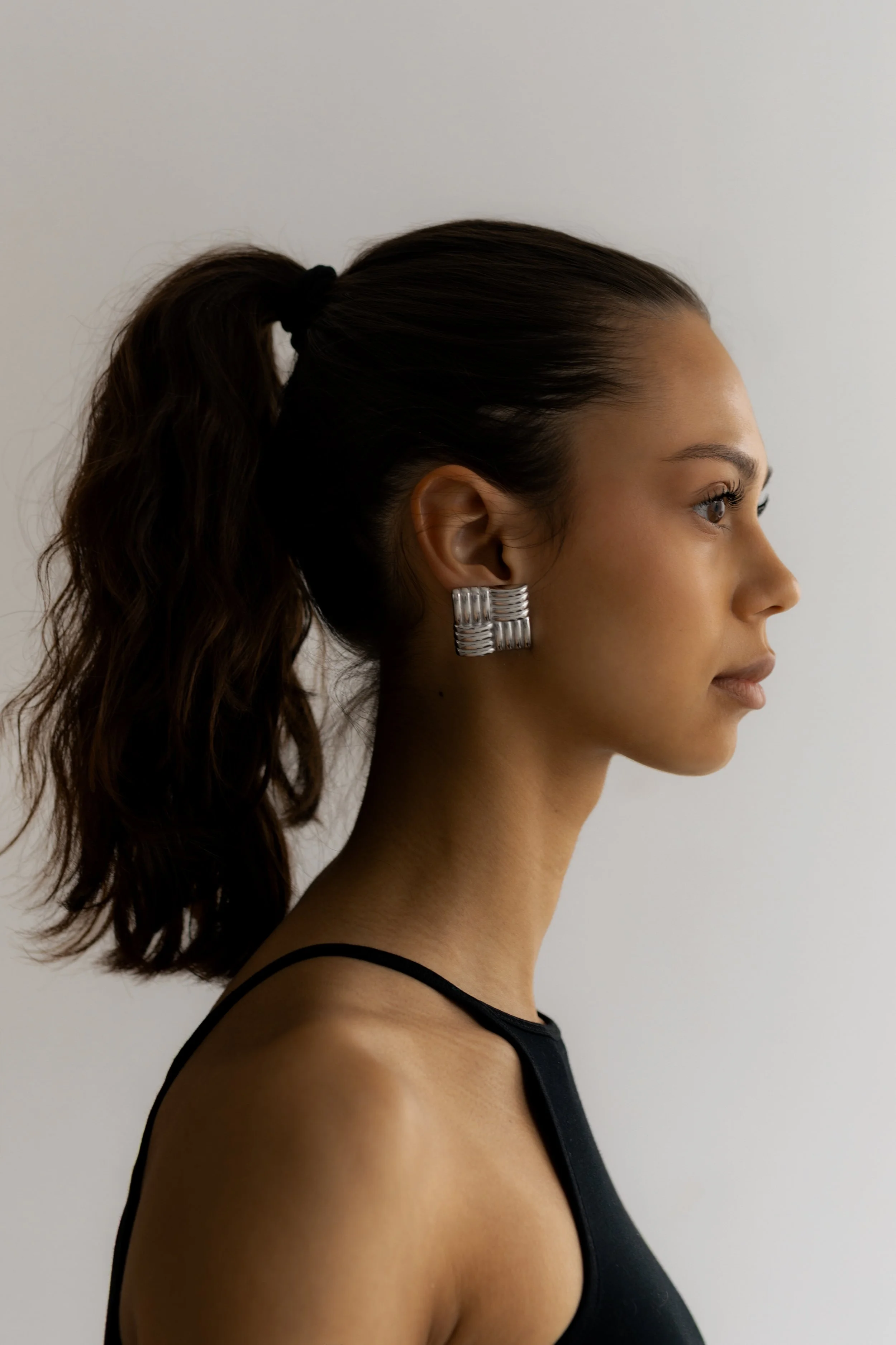 Side profile of a woman with dark brown hair tied in a high ponytail, wearing large silver earrings and a black sleeveless top, against a plain light background.