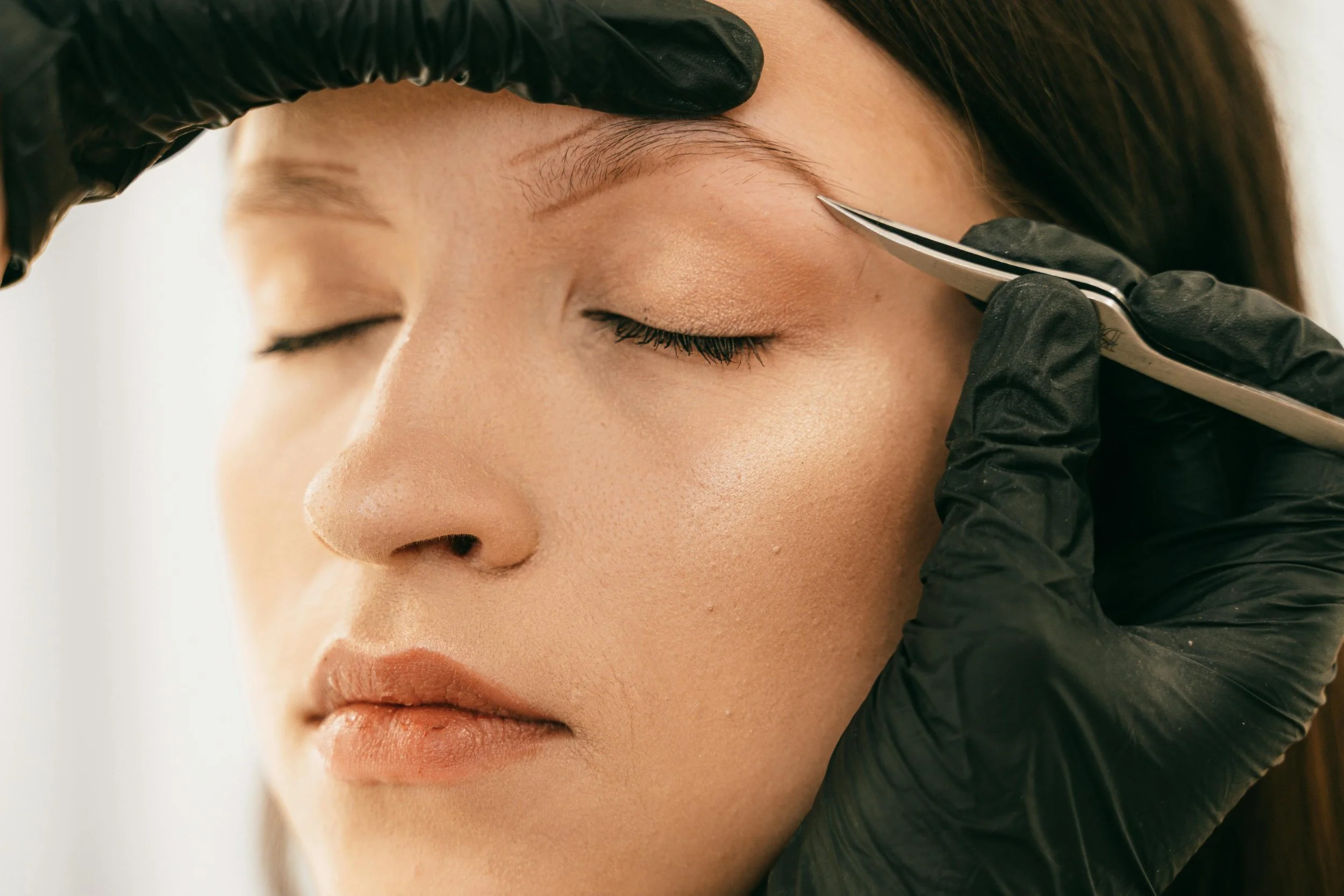 A woman getting her eyebrows shaped with a pair of tweezers by a beautician wearing black gloves.