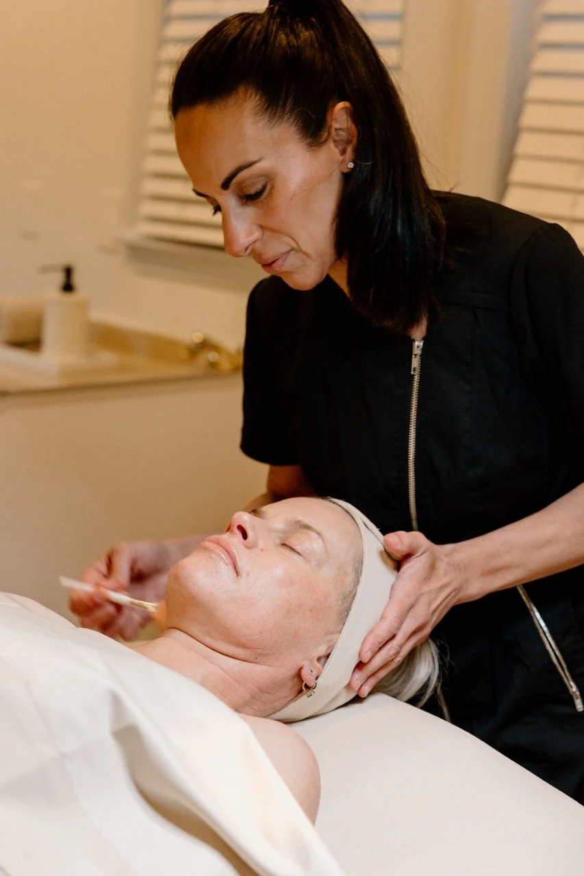 Liz Clapsis Esthetician providing facial treatment to a woman lying on a spa bed, with the woman wearing a headband and the esthetician focused on her work.