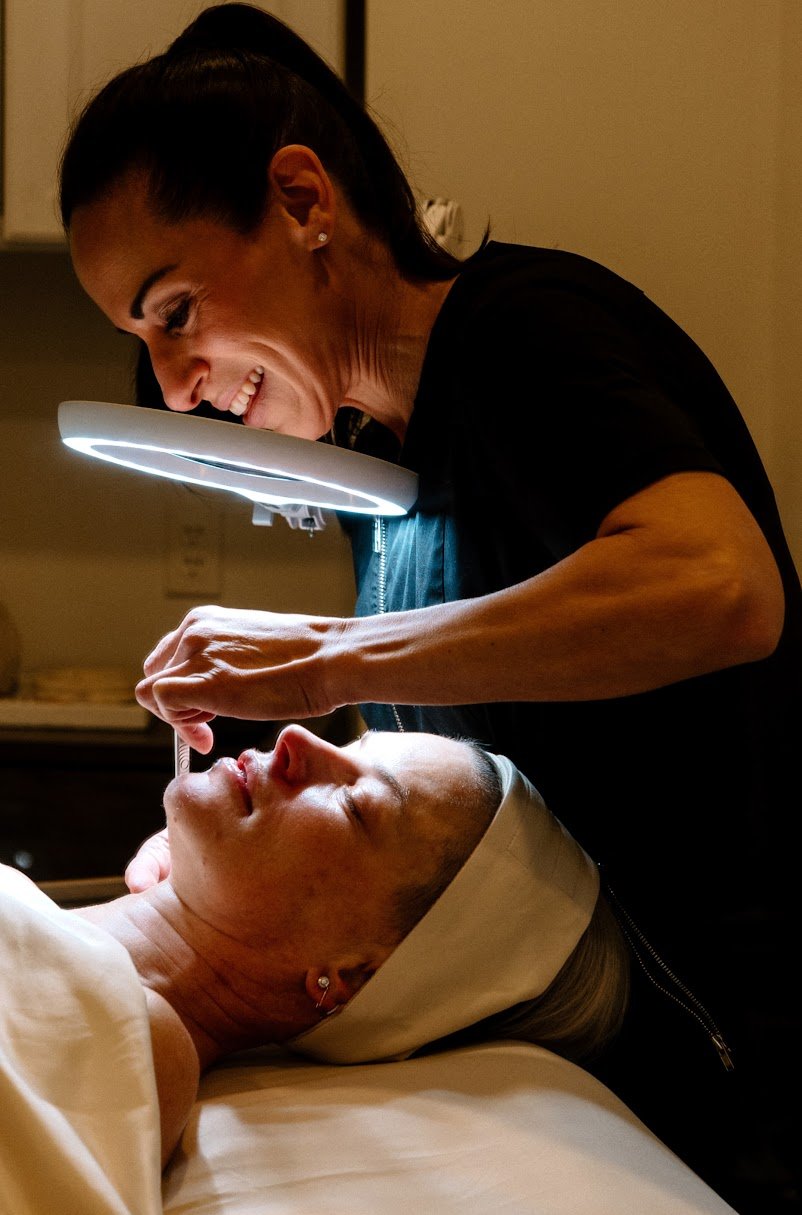 A woman lying in a medical or spa setting while a esthetician Liz Clapsis , examines her face closely with a magnifying light.