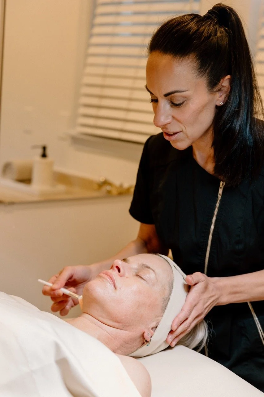 Liz Clapsis giving a skincare treatment to an elderly woman lying on a bed, with her eyes closed, in a room with a window and blinds in the background.