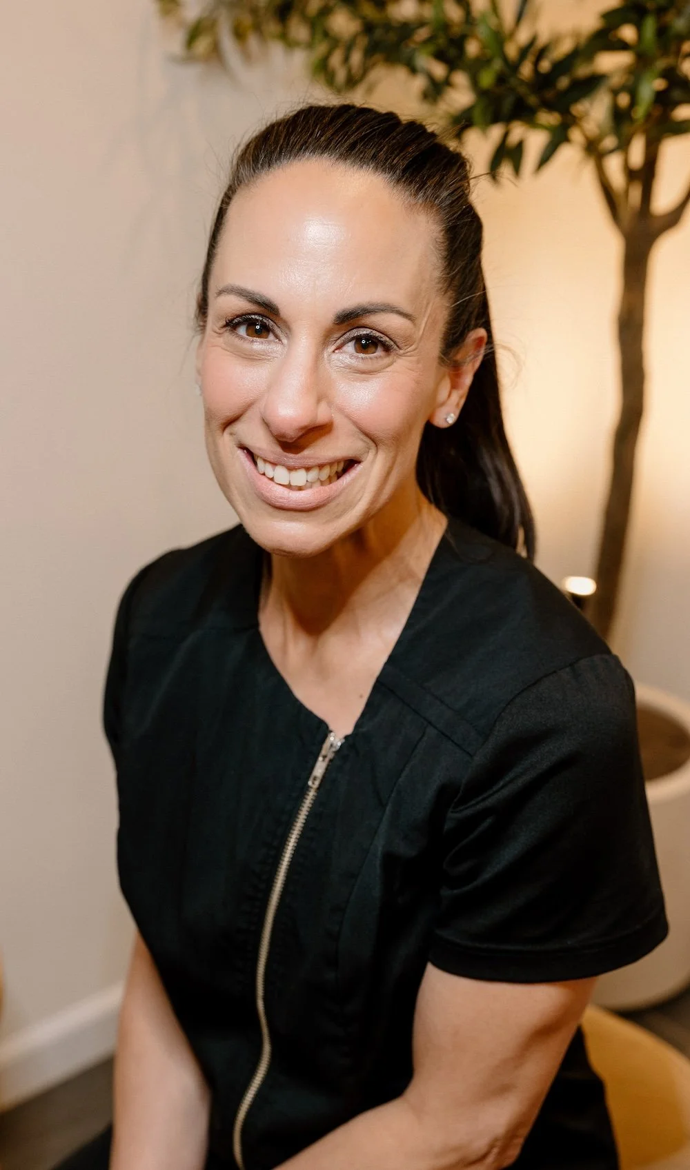 Liz Clapsis with dark hair tied back, smiling, wearing a black top with a zipper, sitting indoors with a potted plant in the background.