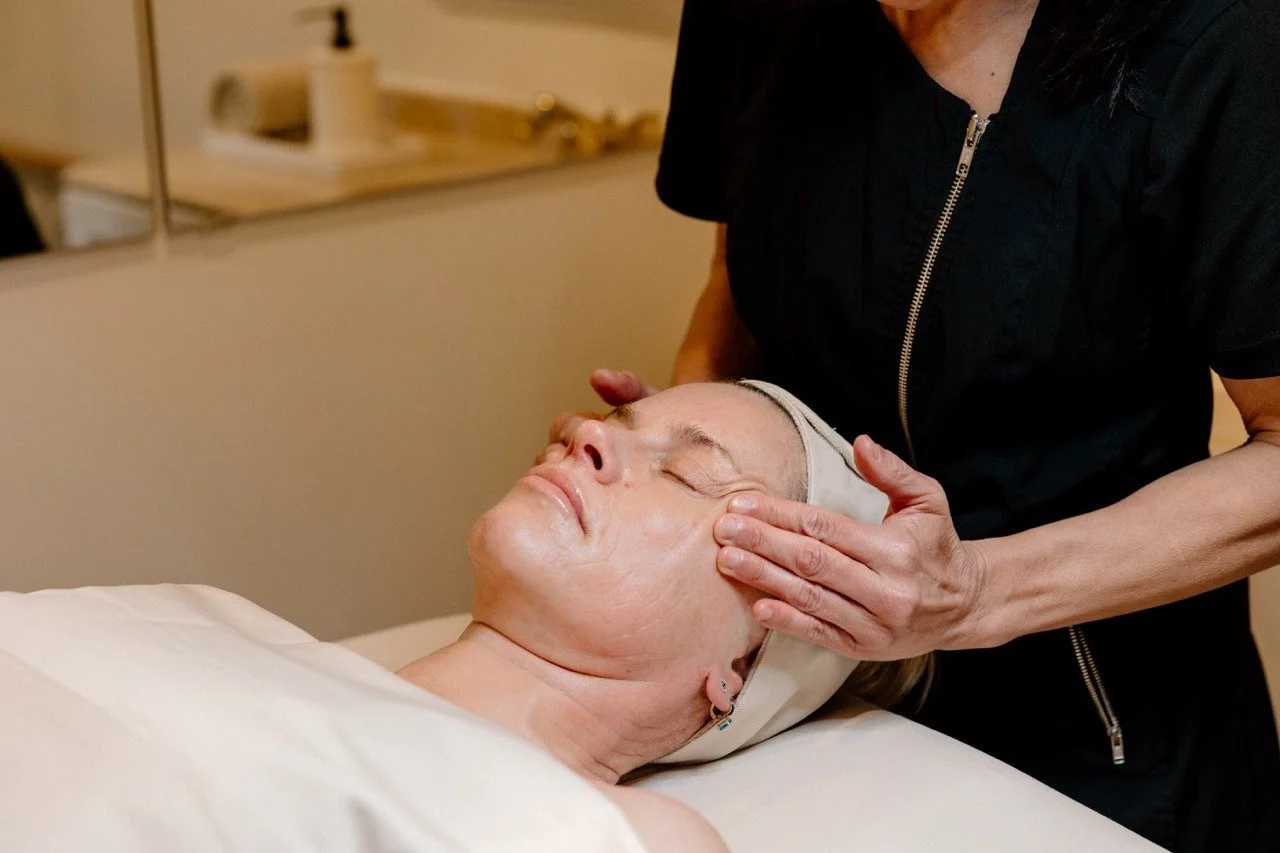 A woman lying on a treatment bed with a towel, receiving a facial massage from Liz Clapsis  in black scrubs in a spa or clinic setting.