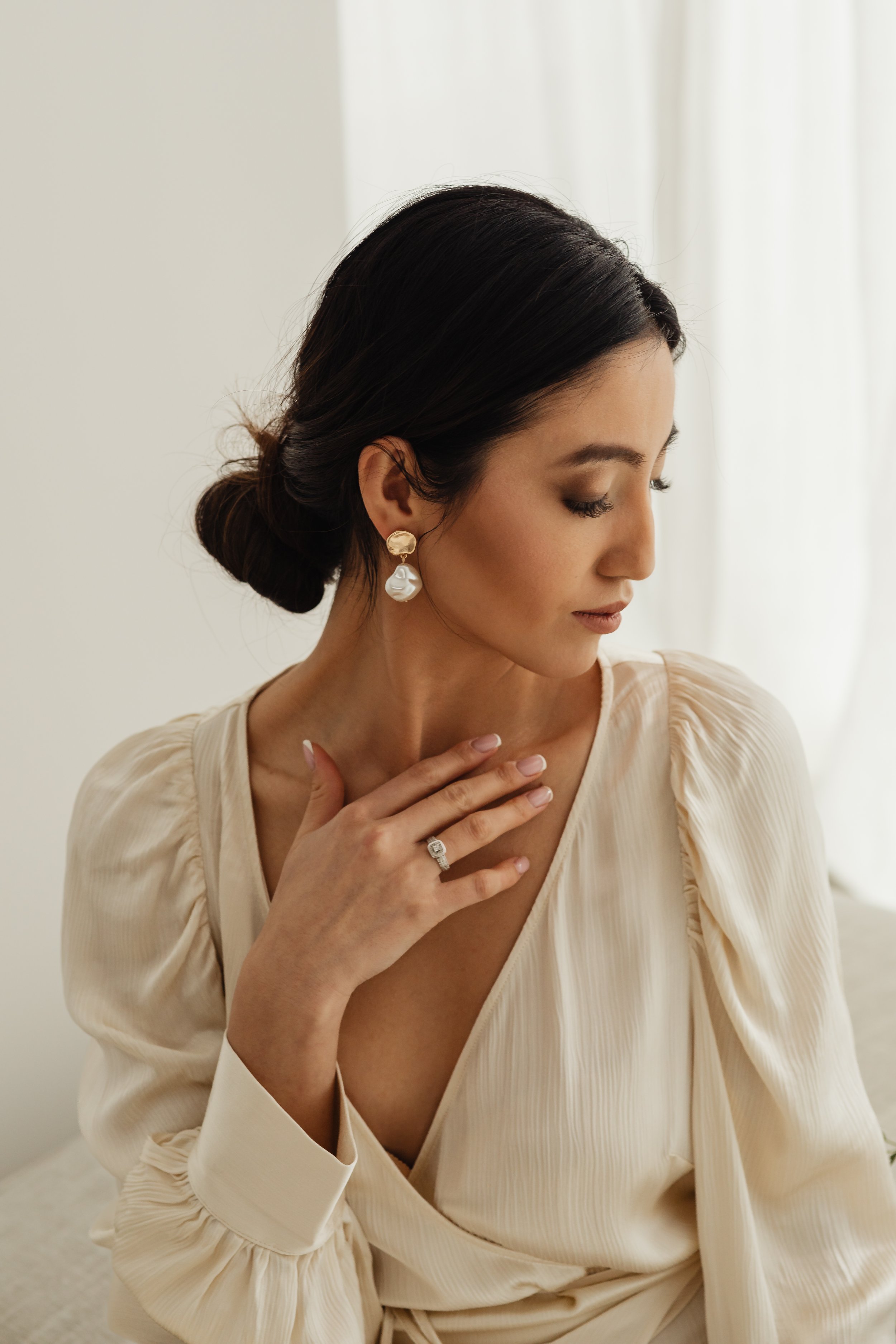 A woman with dark hair in an elegant updo, wearing pearl and gold earrings, a diamond ring, and a cream-colored pleated dress, standing near a window with soft natural light.