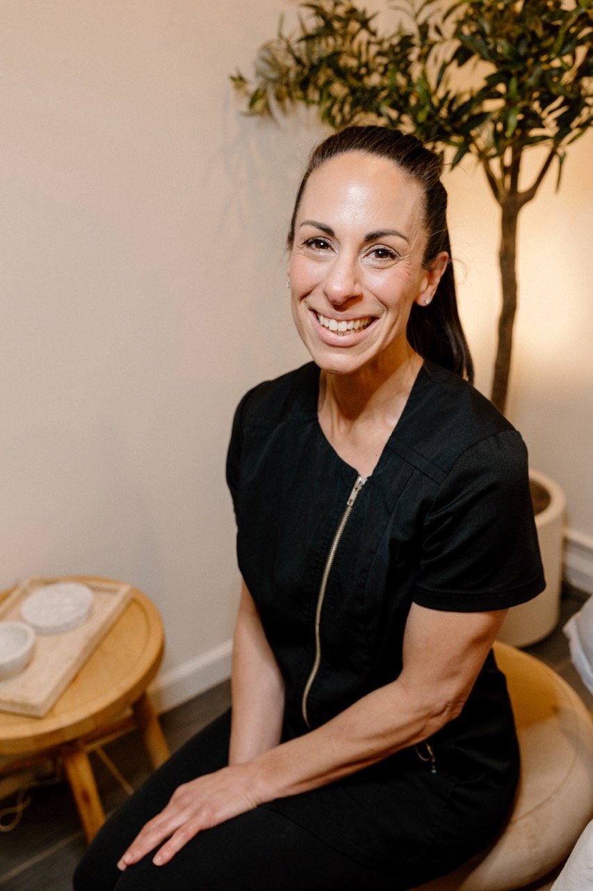 Liz Clapsis with dark hair tied back, wearing a black zip-up top, sitting on a wooden stool in a cozy room with a potted plant and small table with bowls.