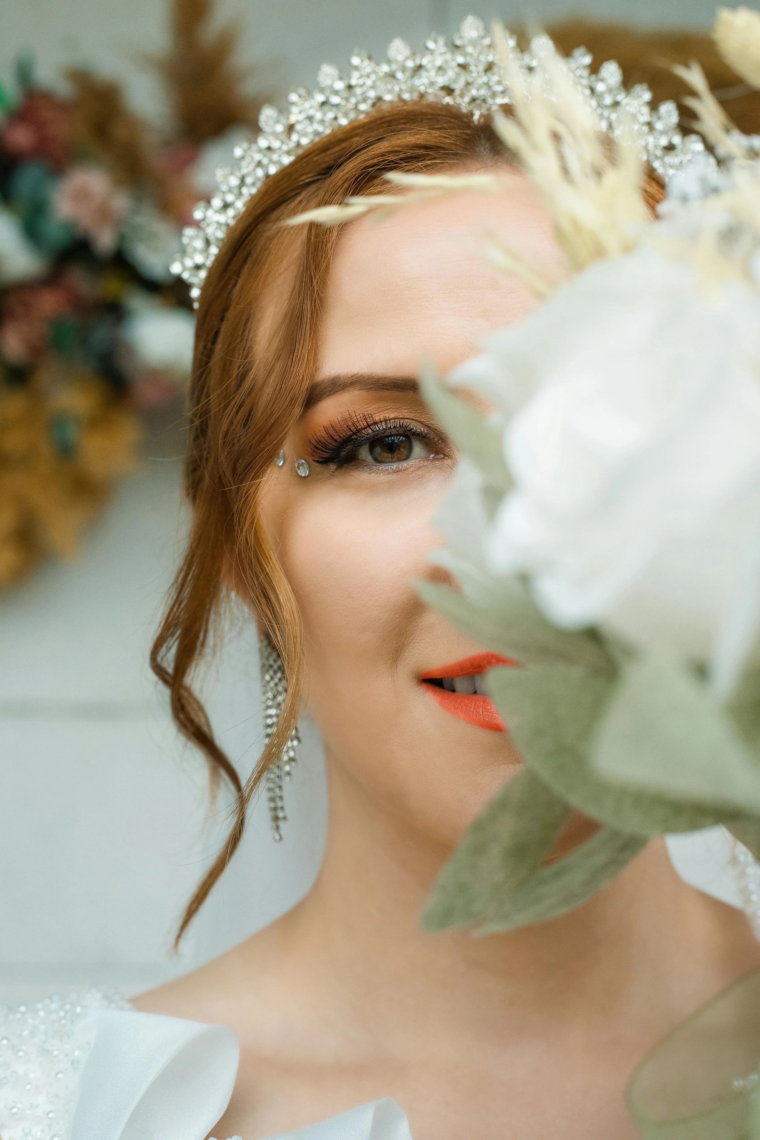 A bride with makeup, jewelry, and a tiara poses partially behind a large white flower.