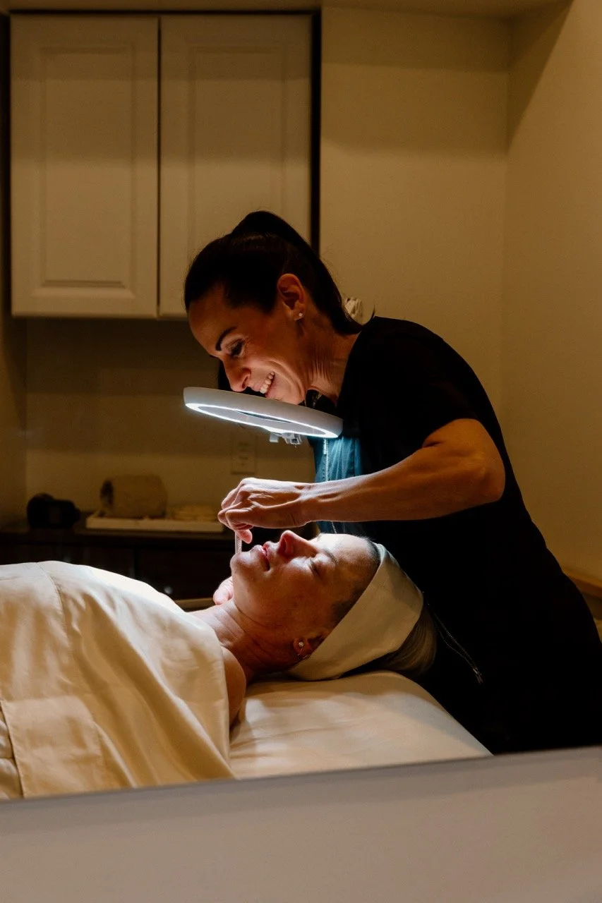 A woman receiving a facial treatment from a professional, Liz Clapsis, in a clinical setting, with the professional using a magnifying light.