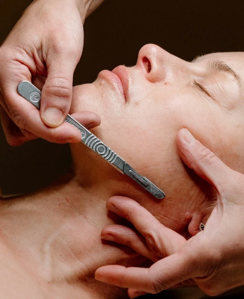 Close-up of a person receiving a facial treatment, with a Liz Clapsis  using a metallic tool on their chin and neck area.