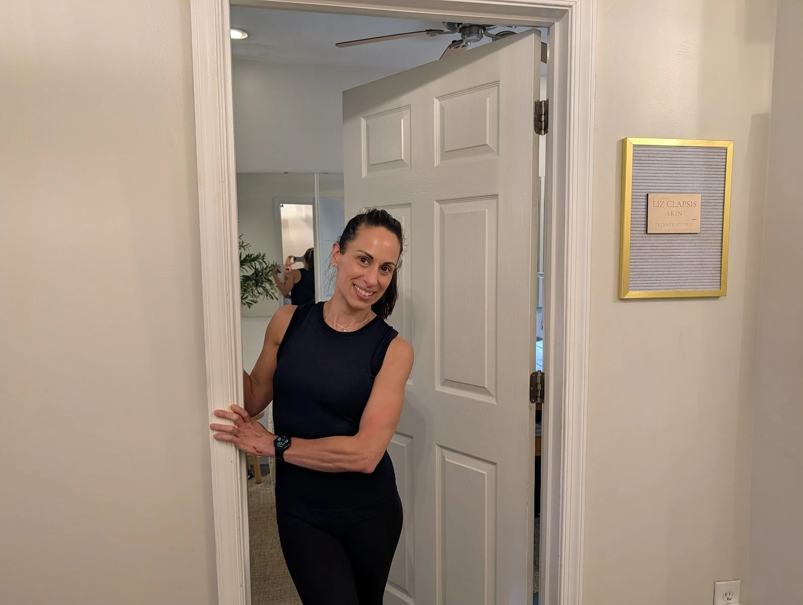 Liz Clapsis in a black tank top and black pants is smiling and peeking out from a doorway, posing for the photograph. Behind her, a woman is taking a picture in a mirror.
