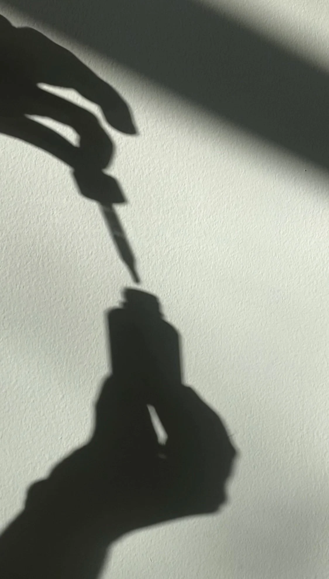 Shadows of a hand holding a small container and a dropper, cast on a light-colored wall.
