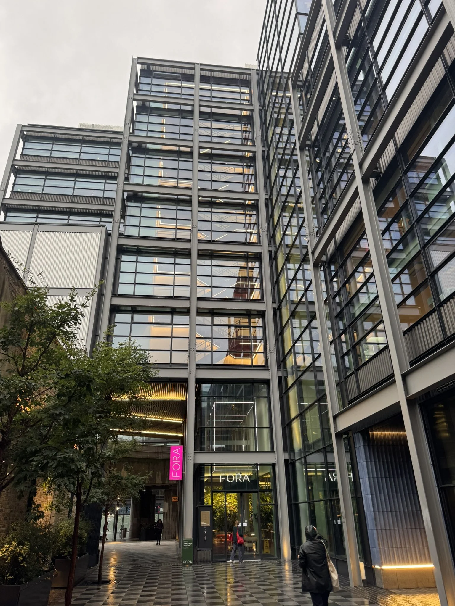 Modern multi-story glass building with a sign that says FORA at the entrance, people walking in and out, and trees on the sidewalk.