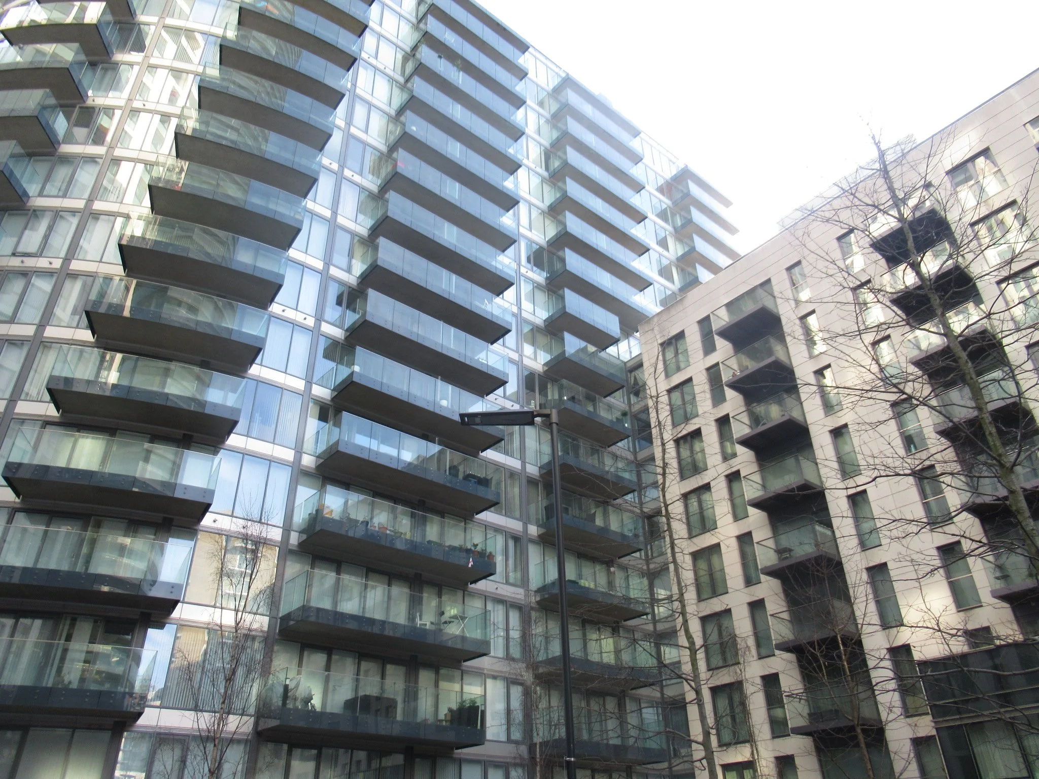 Tall modern glass residential building with multiple balconies, alongside a shorter beige building with windows and balconies, and leafless trees in the foreground.