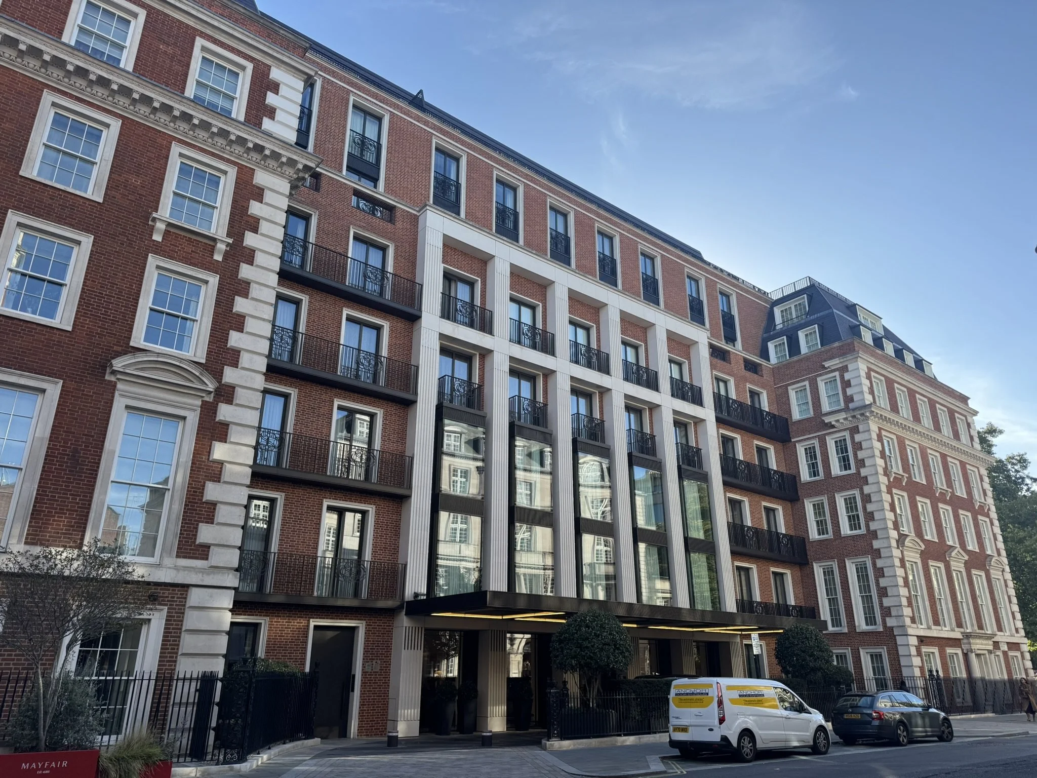 Modern multi-story apartment building with brick and glass facade, black balconies, and a parking area in front with cars.