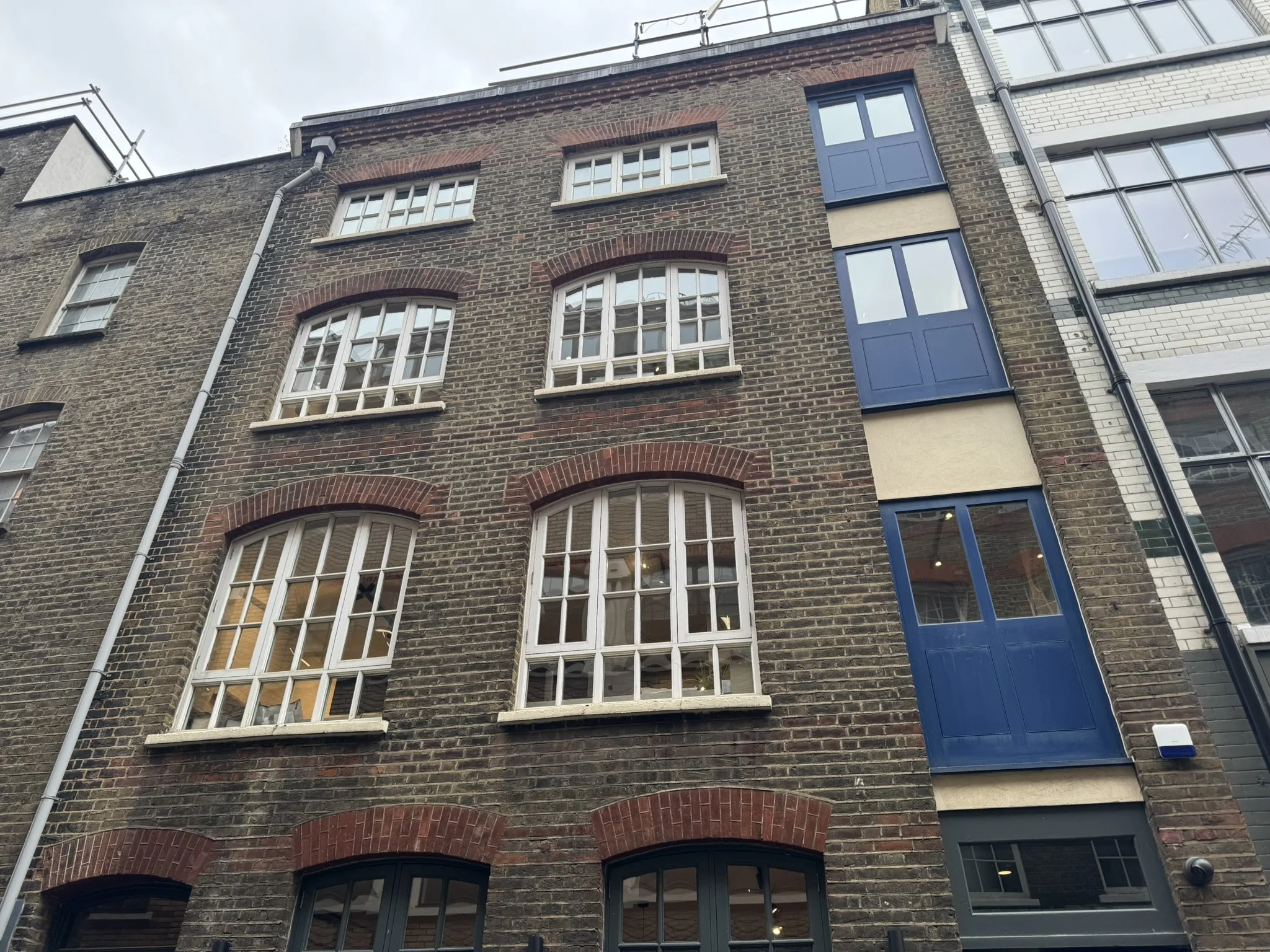 Multi-story brick building with white windowpanes and blue balcony doors, located between older and modern buildings.